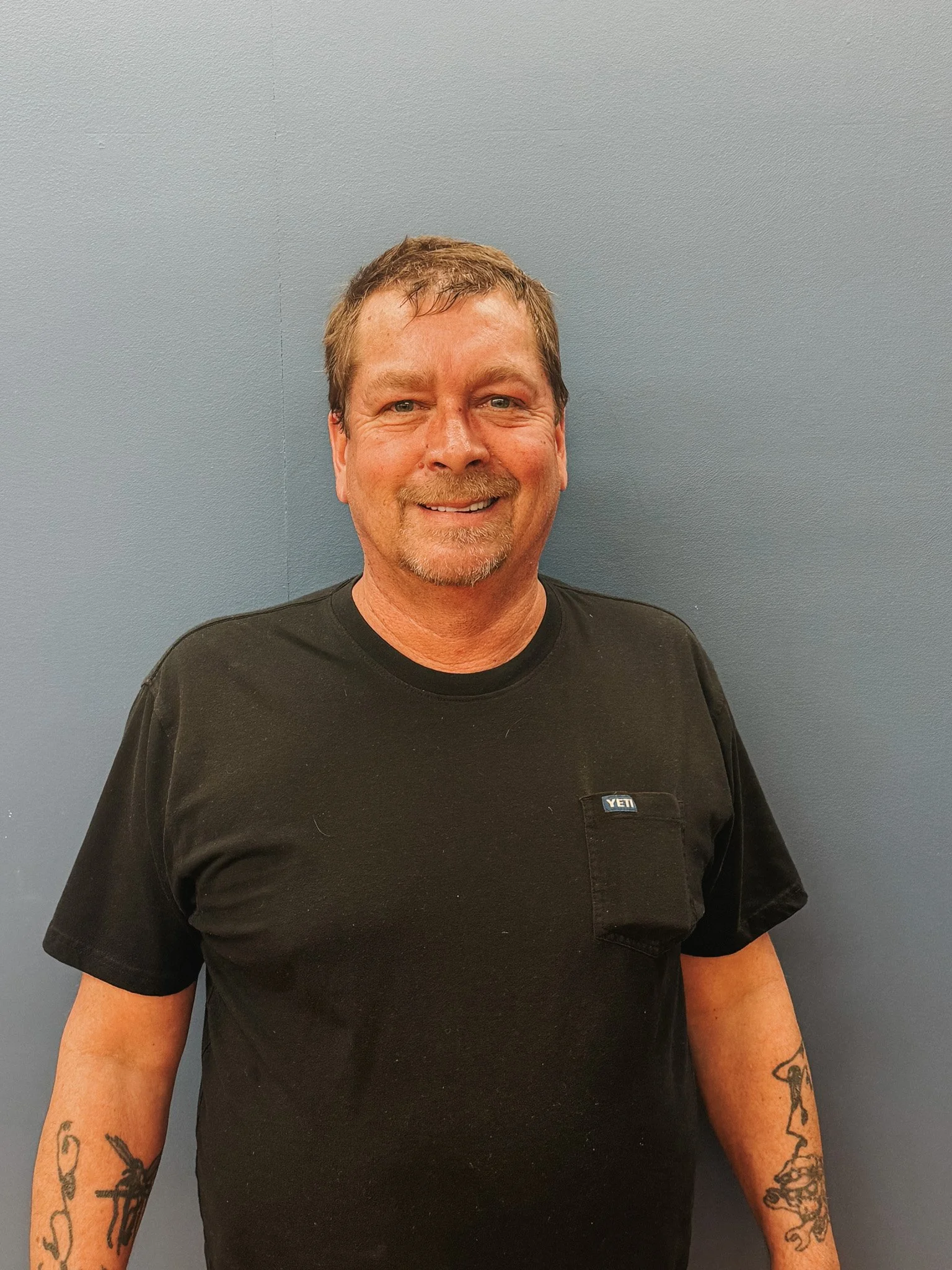A smiling man with brown hair and a goatee, wearing a black T-shirt with a small pocket on the chest, standing against a blue wall.