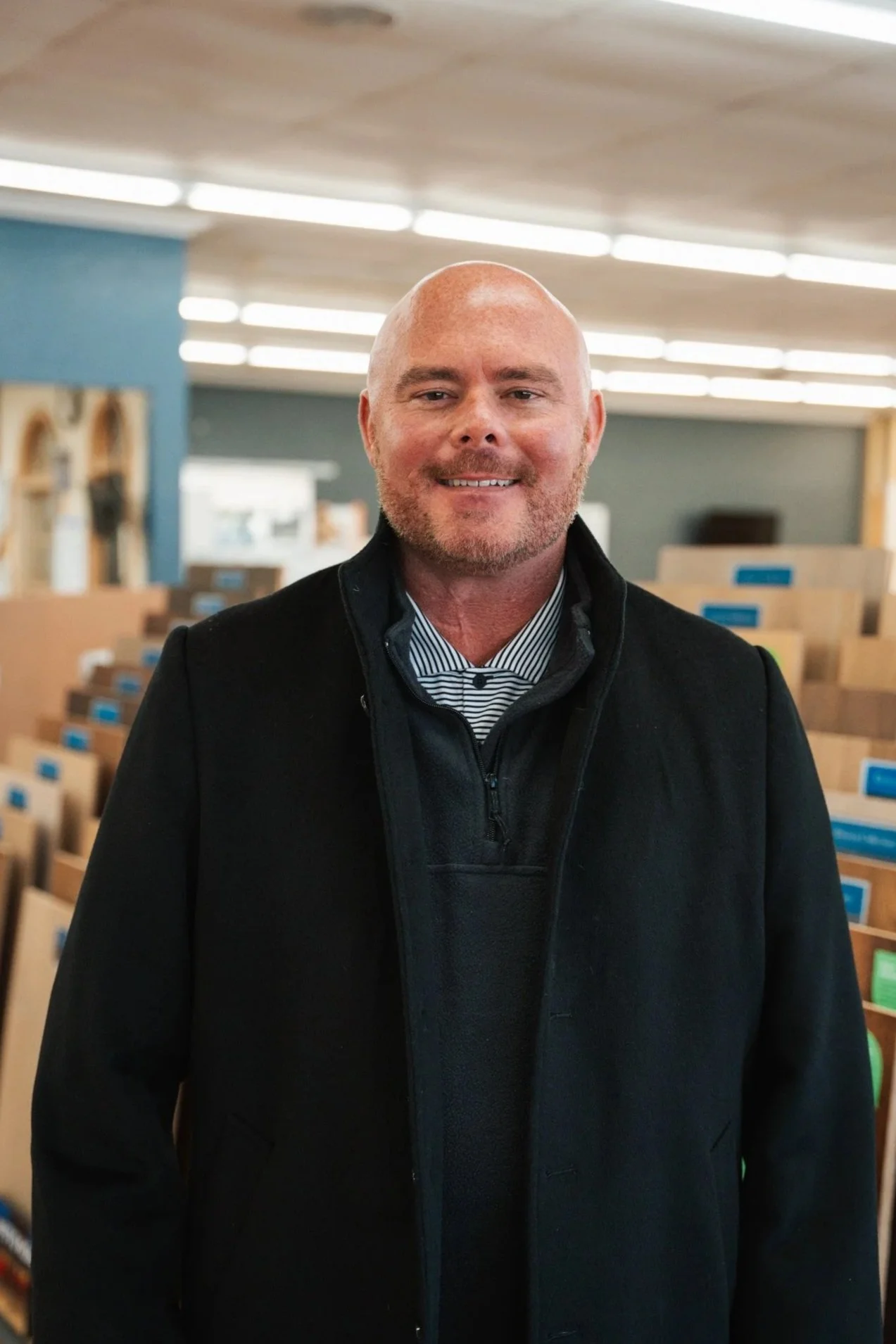 A smiling man with a bald head and beard, wearing a black jacket over a striped shirt, standing indoors in a store with shelves of packages behind him.