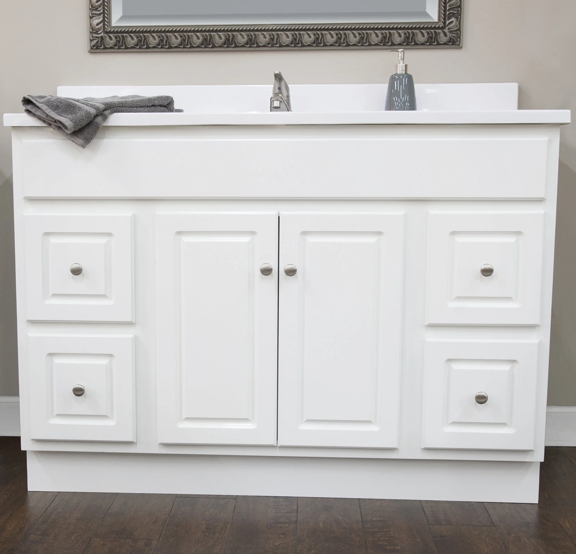White bathroom vanity with towel and soap dispenser, mirror above, dark wooden floor.