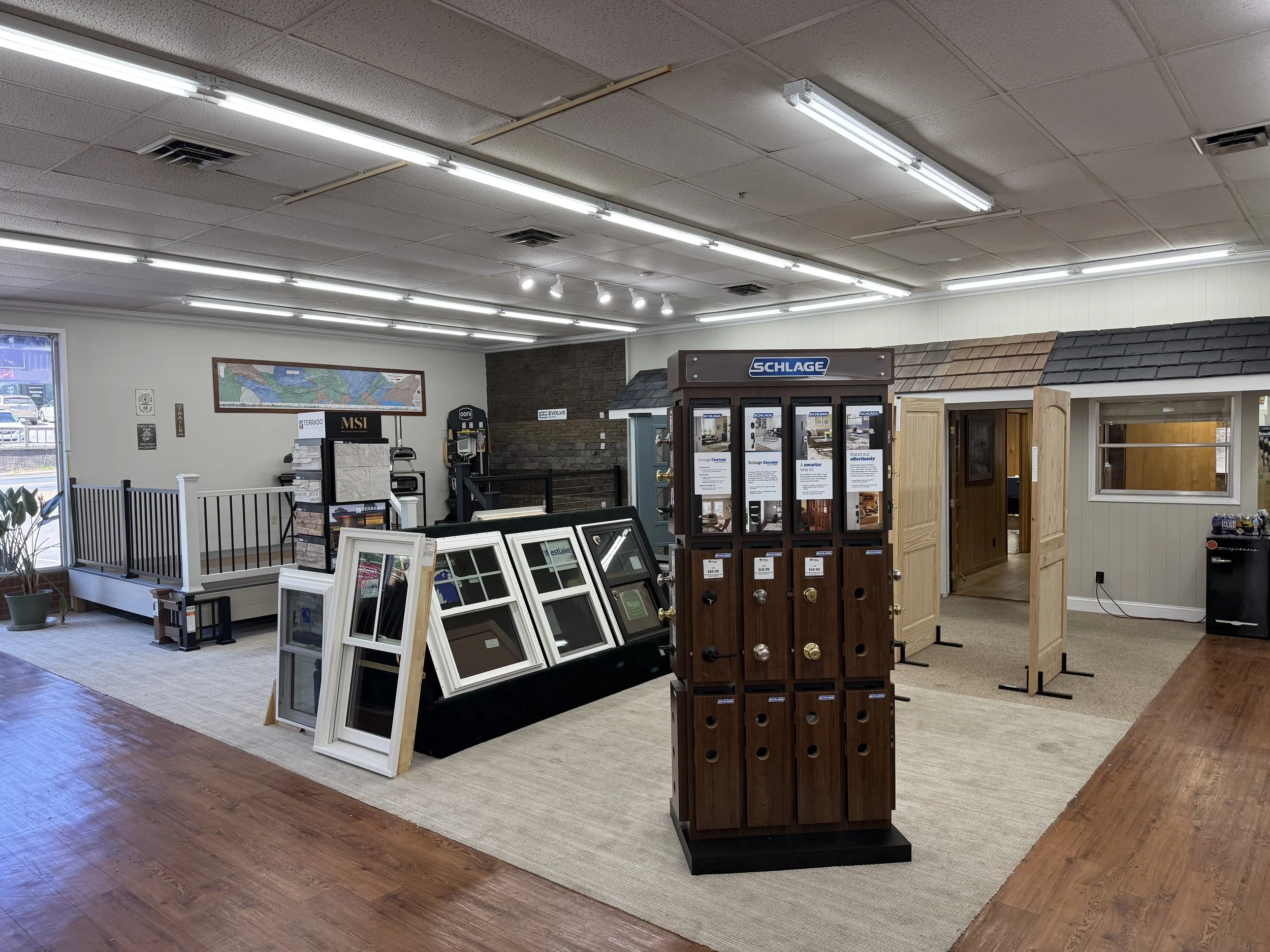 Interior view of a store showing window and door displays, a brick accent wall, and a variety of window frames, blinds, and building materials on display.