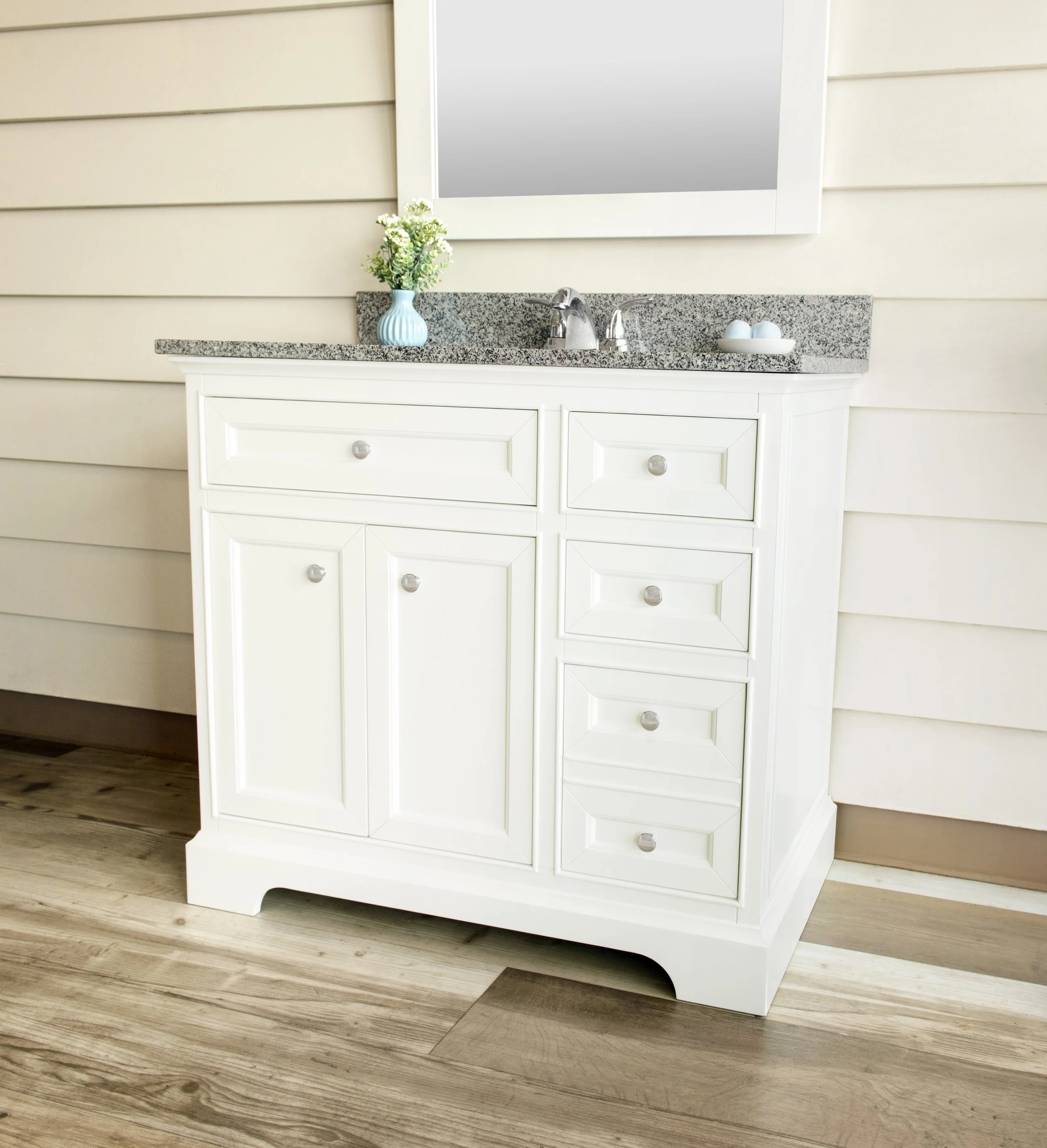 White bathroom vanity with a granite countertop, a single sink with a silver faucet, a small white tray with two blue decorative eggs, and a light blue vase with white flowers, all against a beige wall with horizontal paneling.