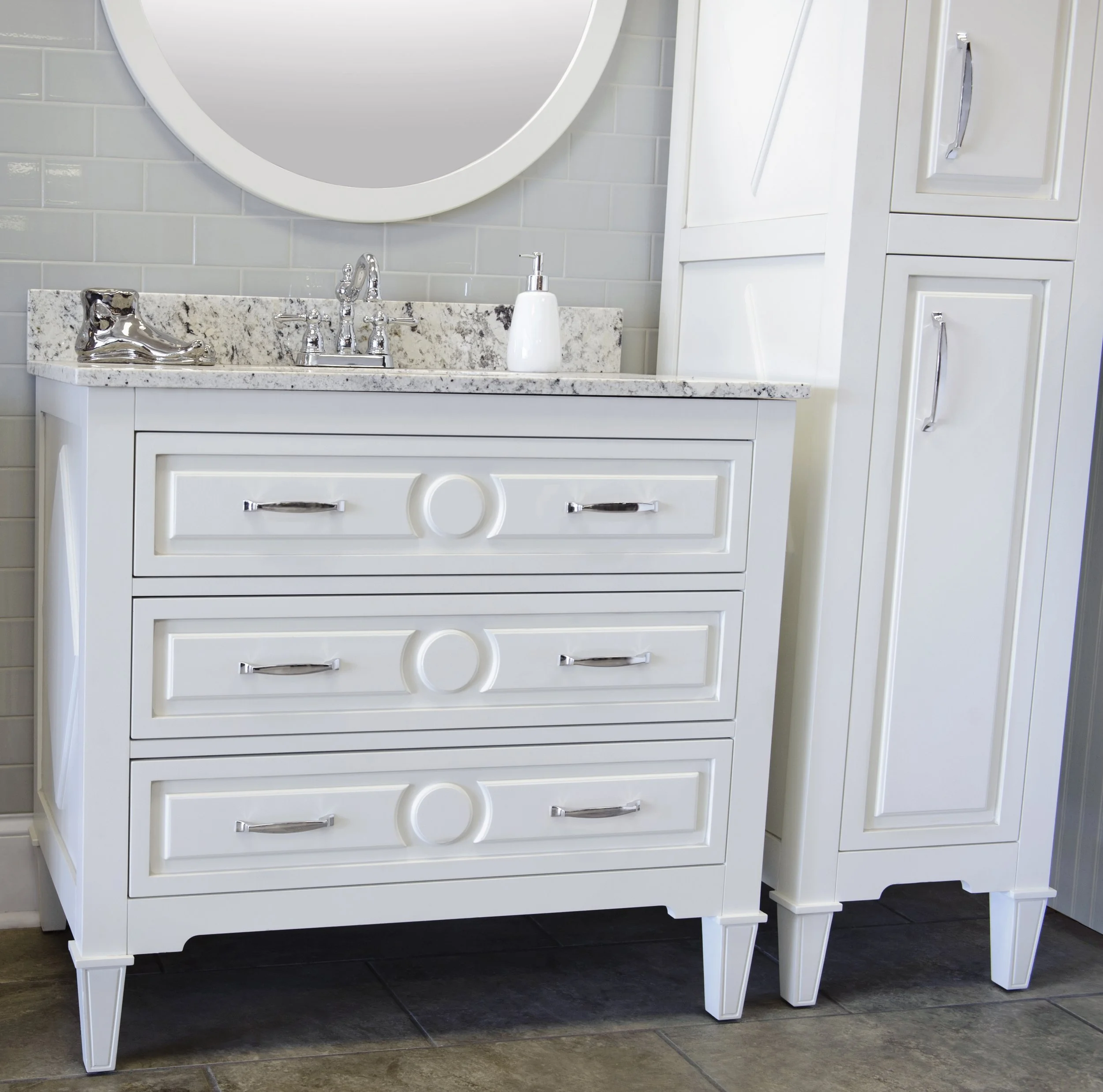 A white bathroom vanity with a granite countertop, a silver decorative shoe, a white liquid soap dispenser, and a silver faucet, with a large oval mirror above and white cabinets to the right.