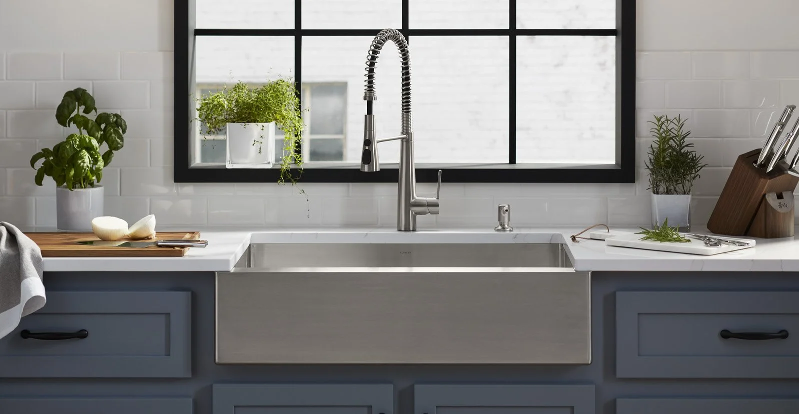 Modern kitchen sink with a stainless steel faucet, plants near window, cutting board with onions, and kitchen utensils on a white countertop.
