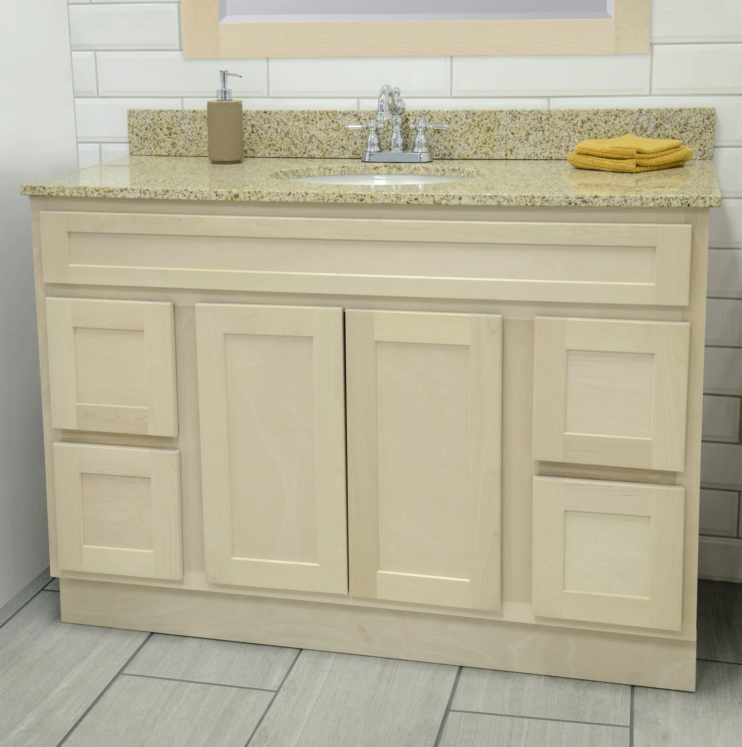 Bathroom sink with a granite countertop, a chrome faucet, a beige soap dispenser, and yellow stacked towels. White tiled wall and light wood cabinetry.