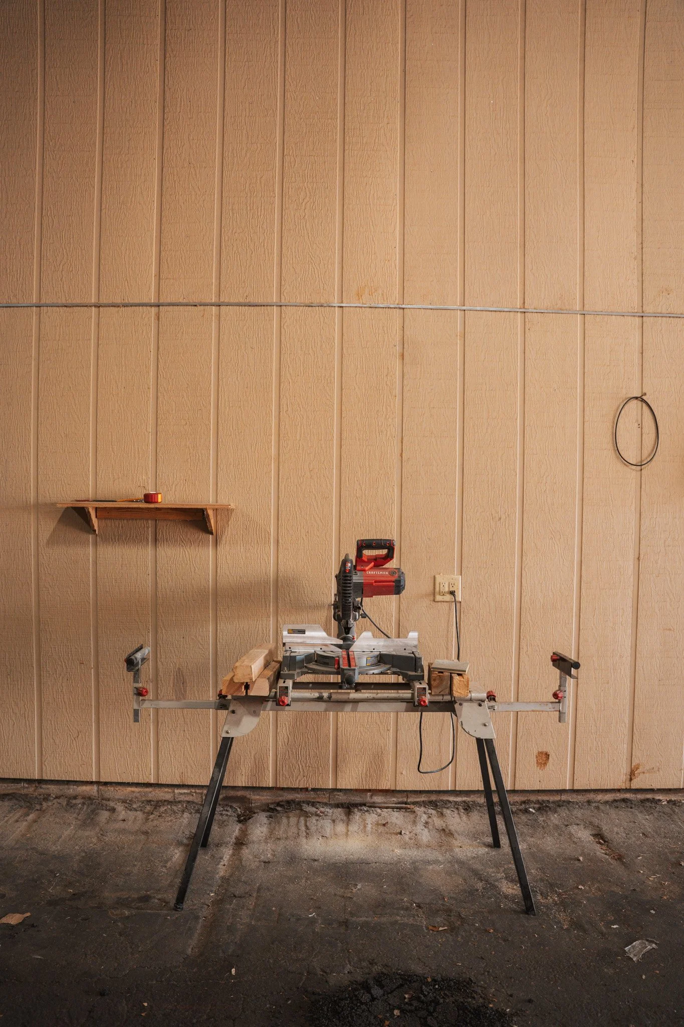 A woodworking miter saw setup with a cordless drill on a wooden stand, against a beige vertical panel wall with a small wooden shelf and a black wire loop hanging.