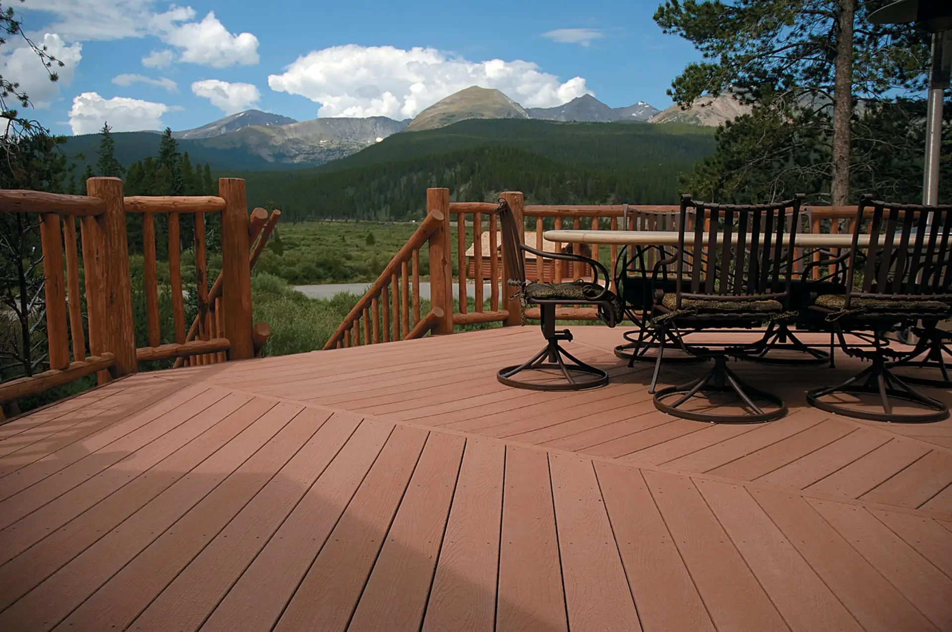 View of a mountain range with green forests on slopes, seen from a wooden deck with patio furniture in the foreground.