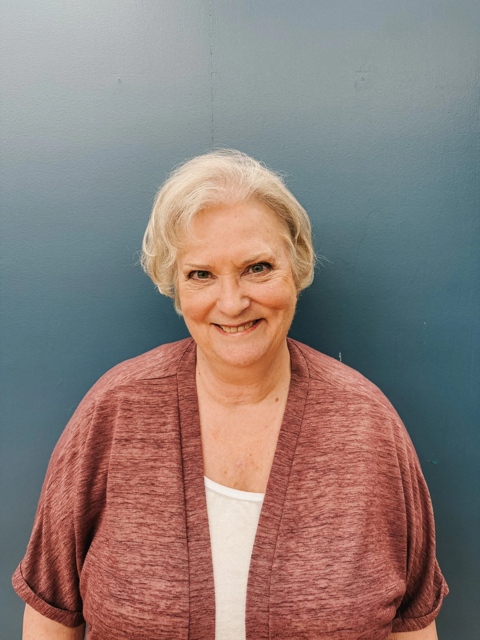 A smiling elderly woman with short, wavy blonde hair, wearing a white top and a mauve cardigan, standing against a blue wall.