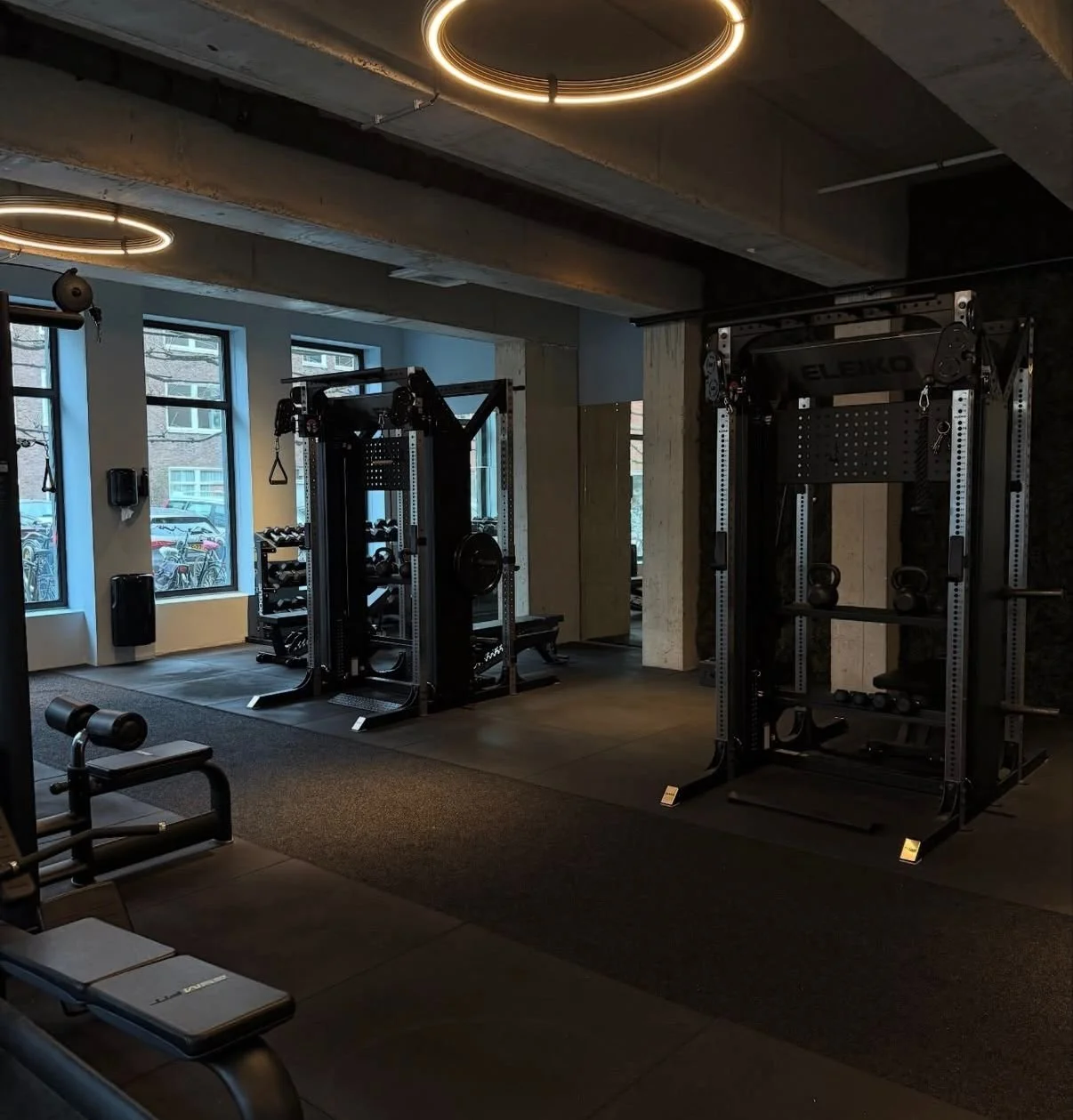 Empty modern gym with large windows, fitness equipment including cable machines, kettlebells, dumbbells, and a workout bench, illuminated by circular ceiling lights.