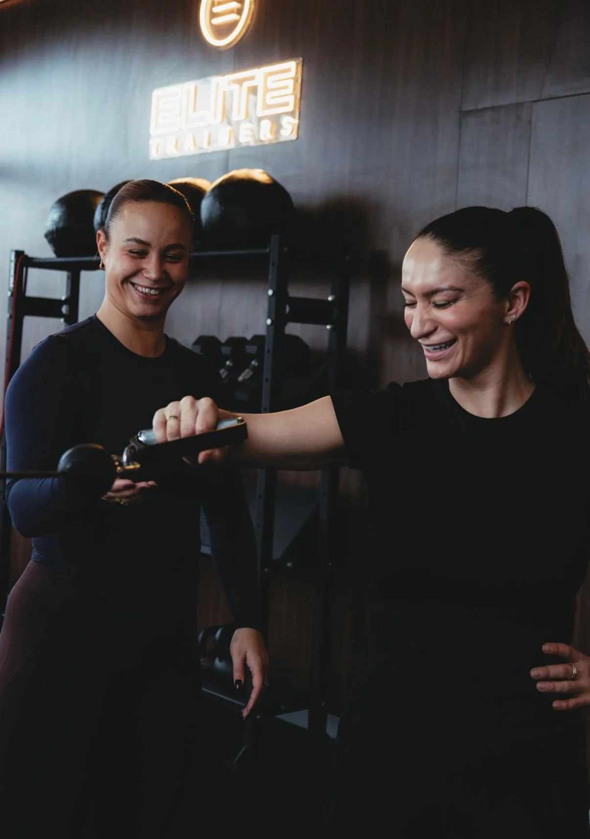 Two women in a gym, one assisting the other with a piece of exercise equipment, smiling.