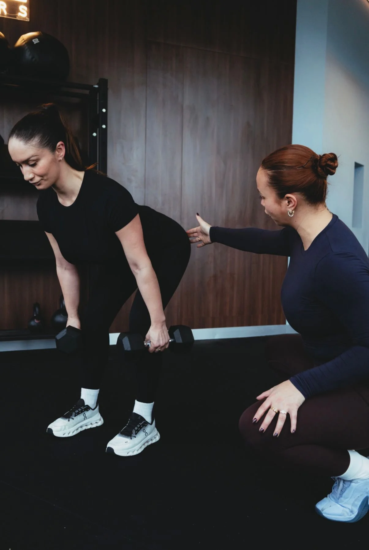 A woman demonstrating a workout exercise with dumbbells while a trainer guides her in a gym with wood-paneled walls and exercise equipment.