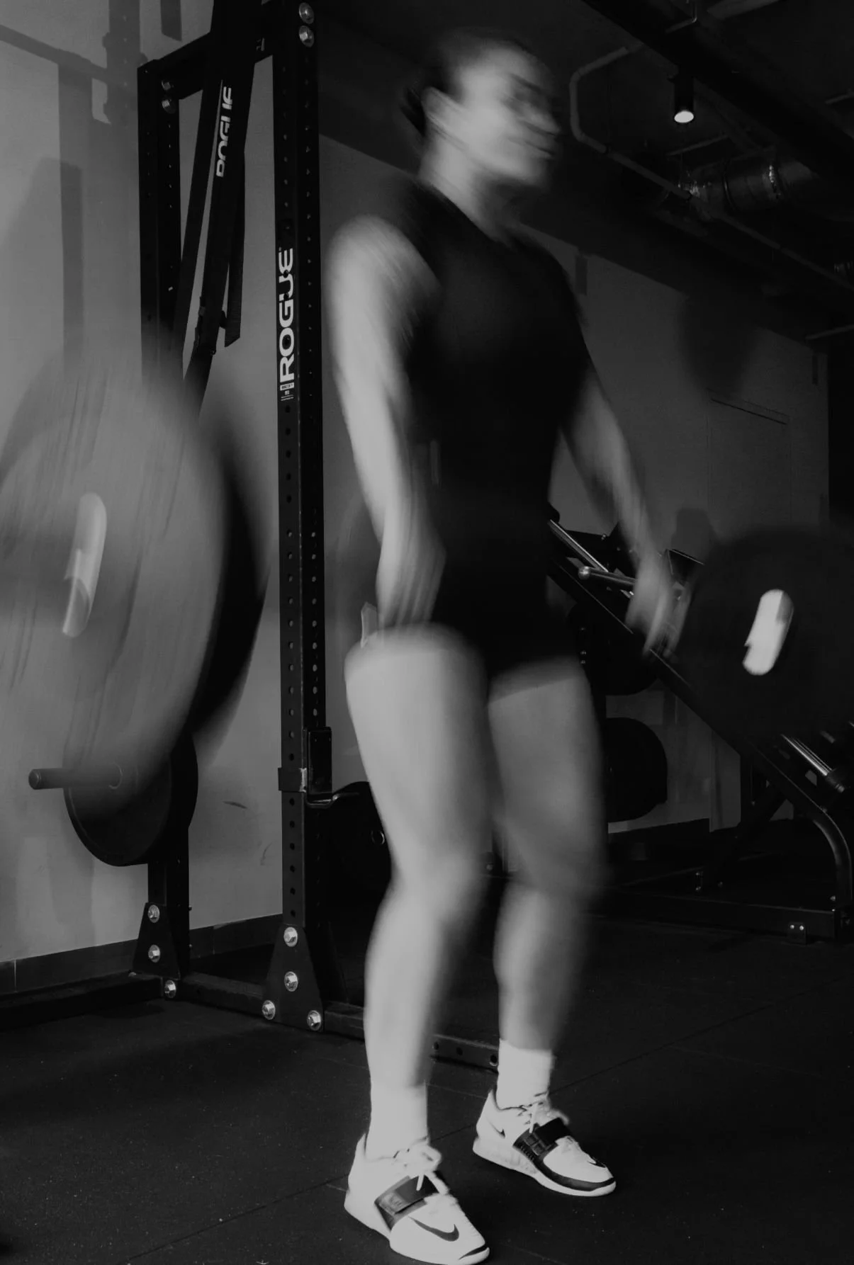 A person lifting weights in a gym, blurred due to motion, wearing athletic shorts, tank top, and sneakers, with gym equipment visible in the background.