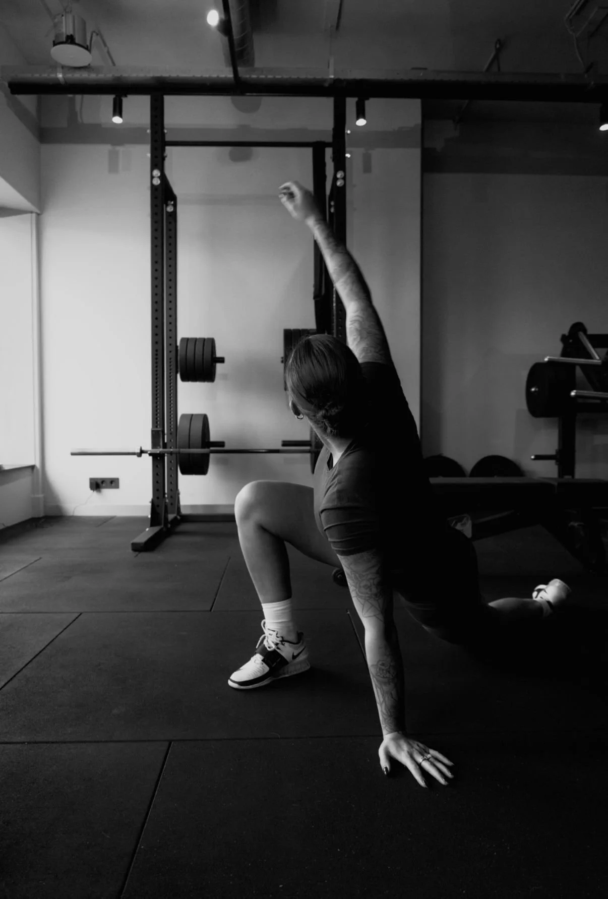 A woman exercising on the floor of a gym, stretching with one hand extended overhead and the other hand on the ground, near weightlifting equipment.