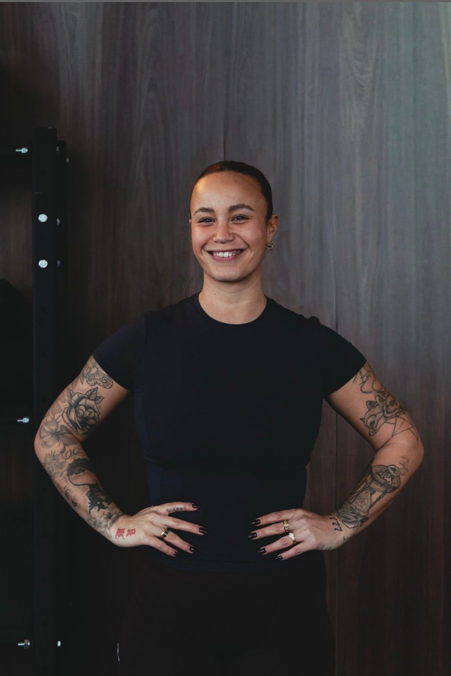A smiling woman with tattoos on her arms, short hair, and wearing a black t-shirt, standing with hands on hips in front of a dark wood-paneled wall.