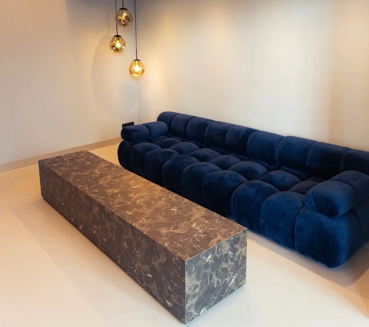 Modern living room with a plush, dark blue velvet sectional sofa, a long marble coffee table, and hanging pendant lights with warm glow in the background.