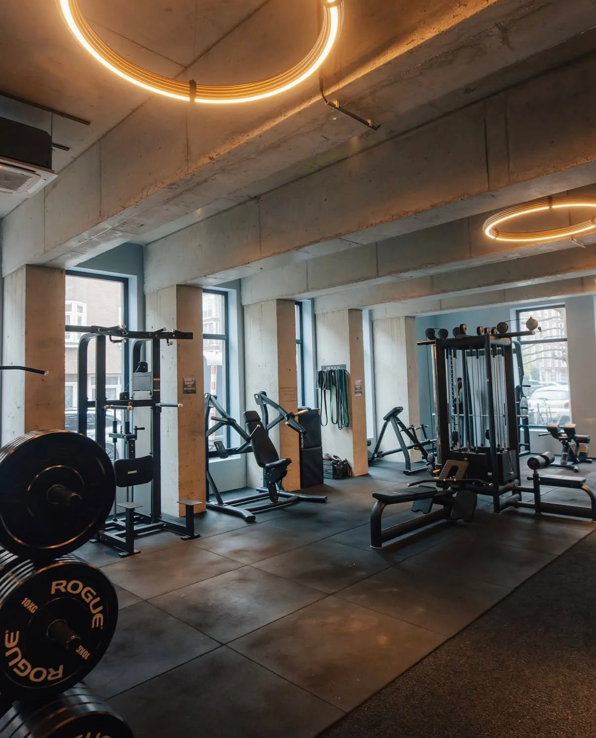 Interior of a modern gym with various exercise equipment including weights, cardio machines, and resistance training stations, illuminated by circular overhead lights and natural light from large windows.