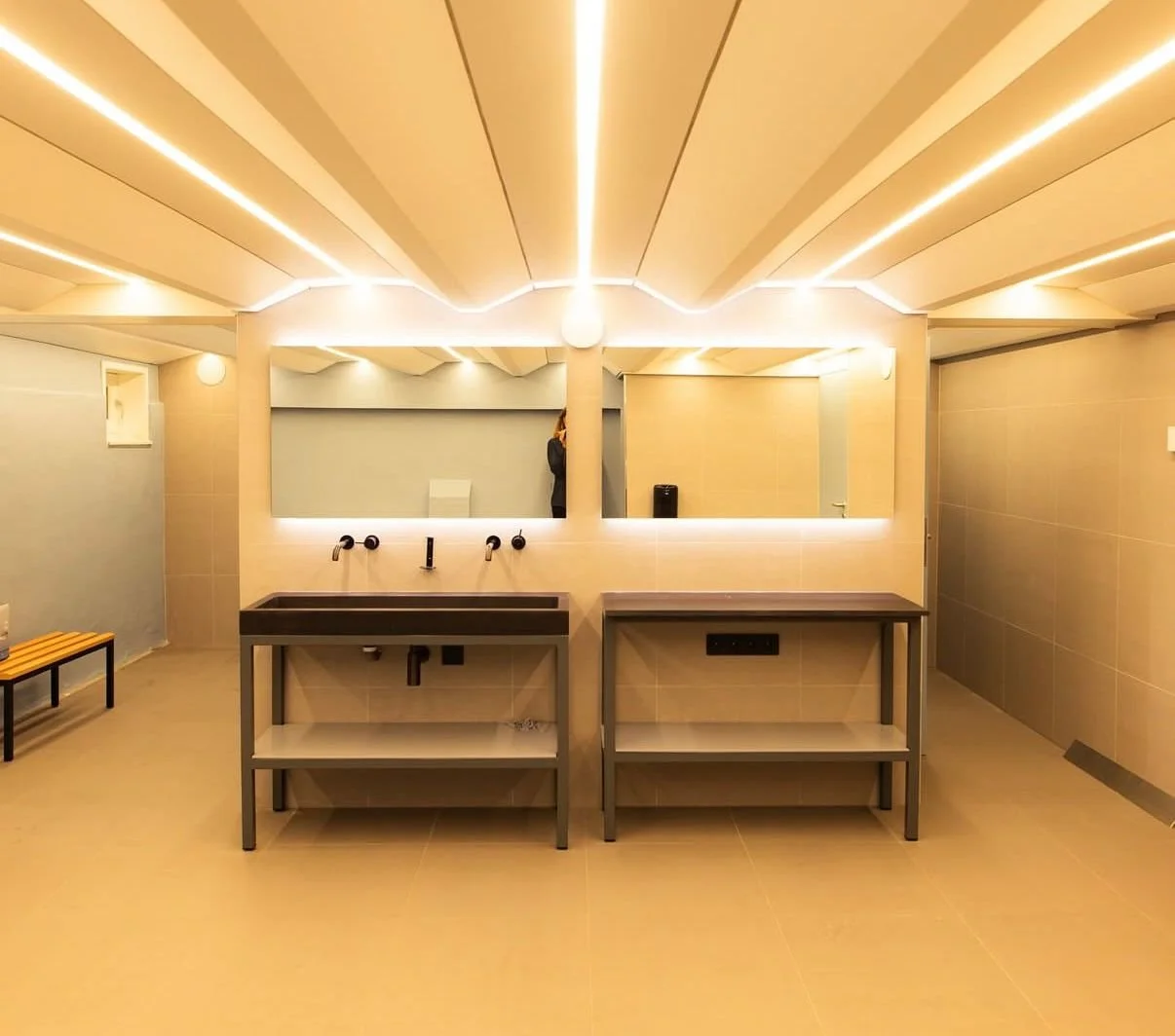 Modern, minimalistic bathroom with two stainless steel sinks, large mirror, and beige tiled walls and floor, illuminated with ceiling lights.
