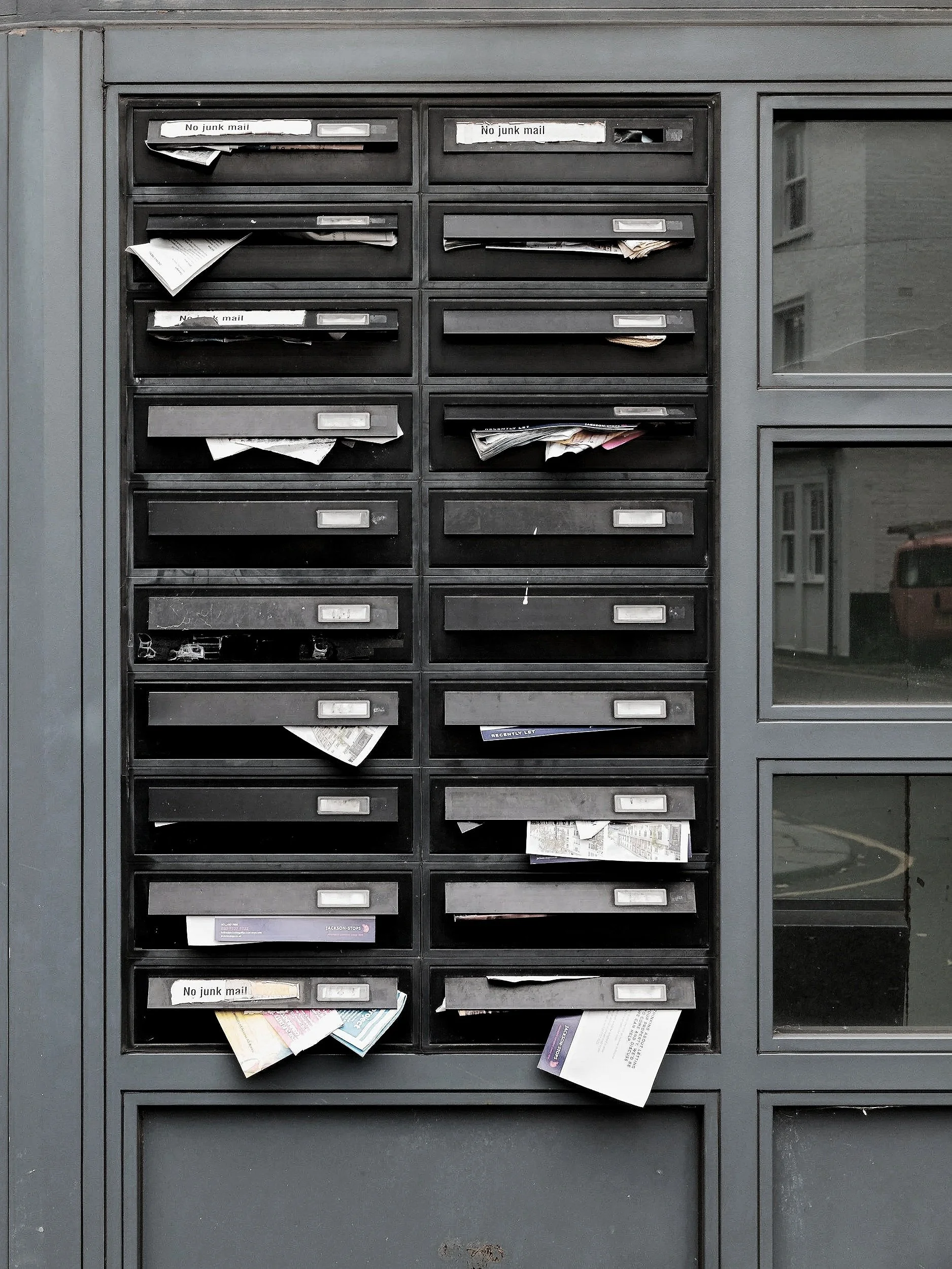 Black mailboxes filled with scattered and overflowing mail, some labeled 'No junk mail,' in an apartment building lobby.
