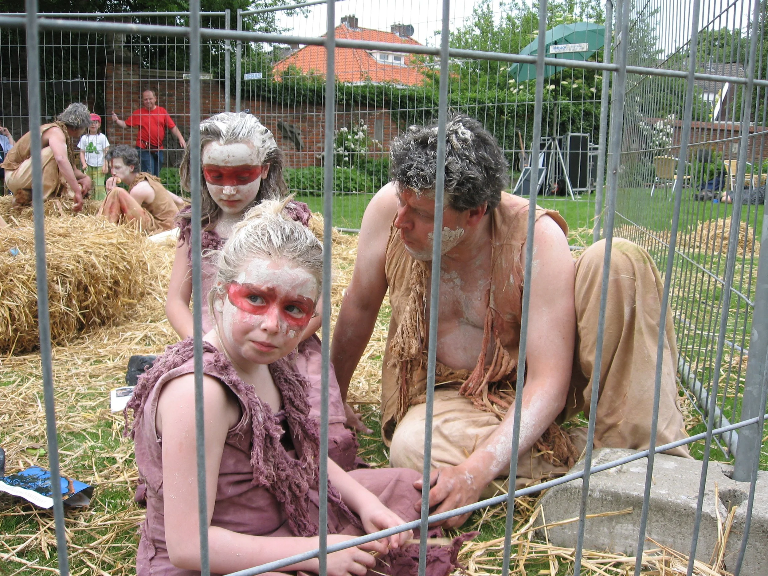 Mensen in verkleedkleding en make-up die zich verkleden als oermensen, in een omheinde ruimte met stro en gras, mogelijk tijdens een livetheatervoorstelling of festival
