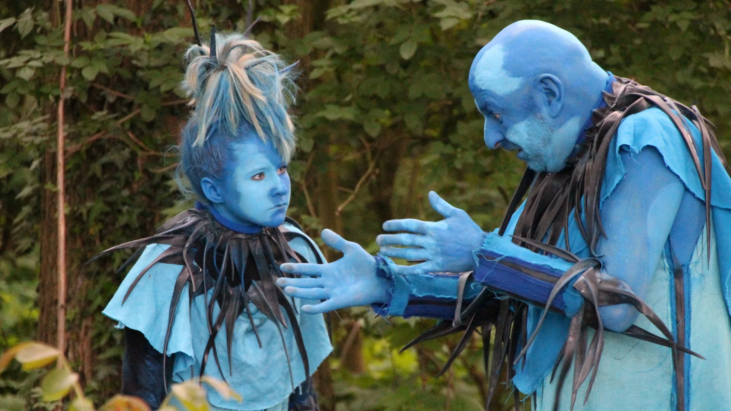 Twee mensen gekleed als blauwe fantasie-personages met blauwe gezichtsbeschildering en kostuums met zwarte en blauwe bladeren, in een bos.