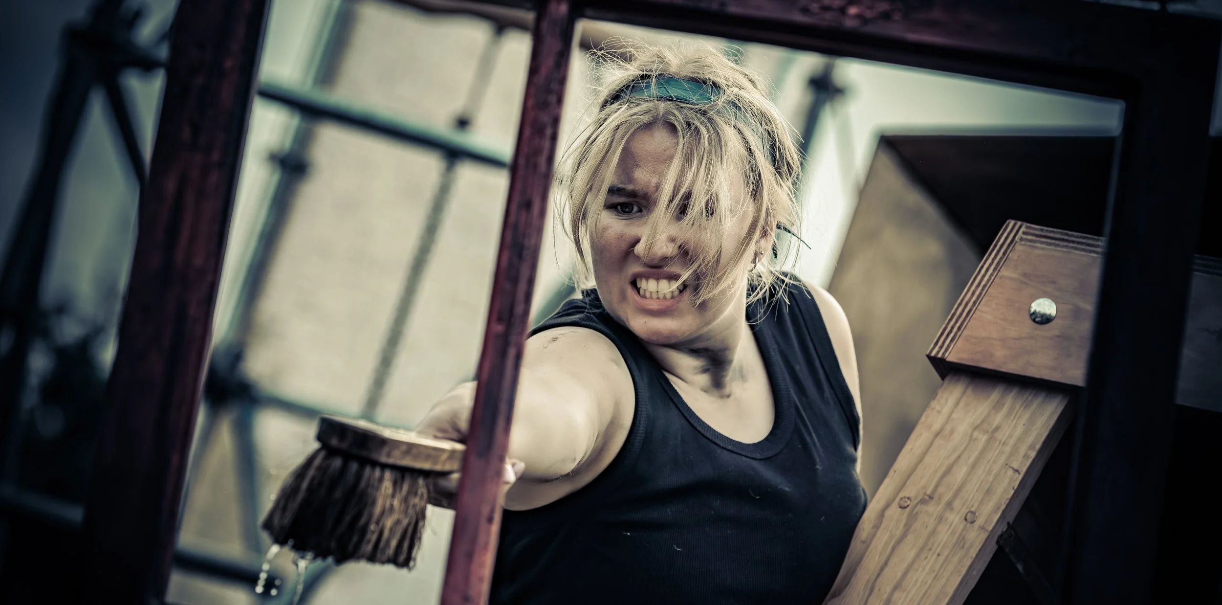 Een vrouw met een vlekkerig gezicht en warrig blond haar die agressief met haar vuist uit een raam slaat in een soort werkomgeving.