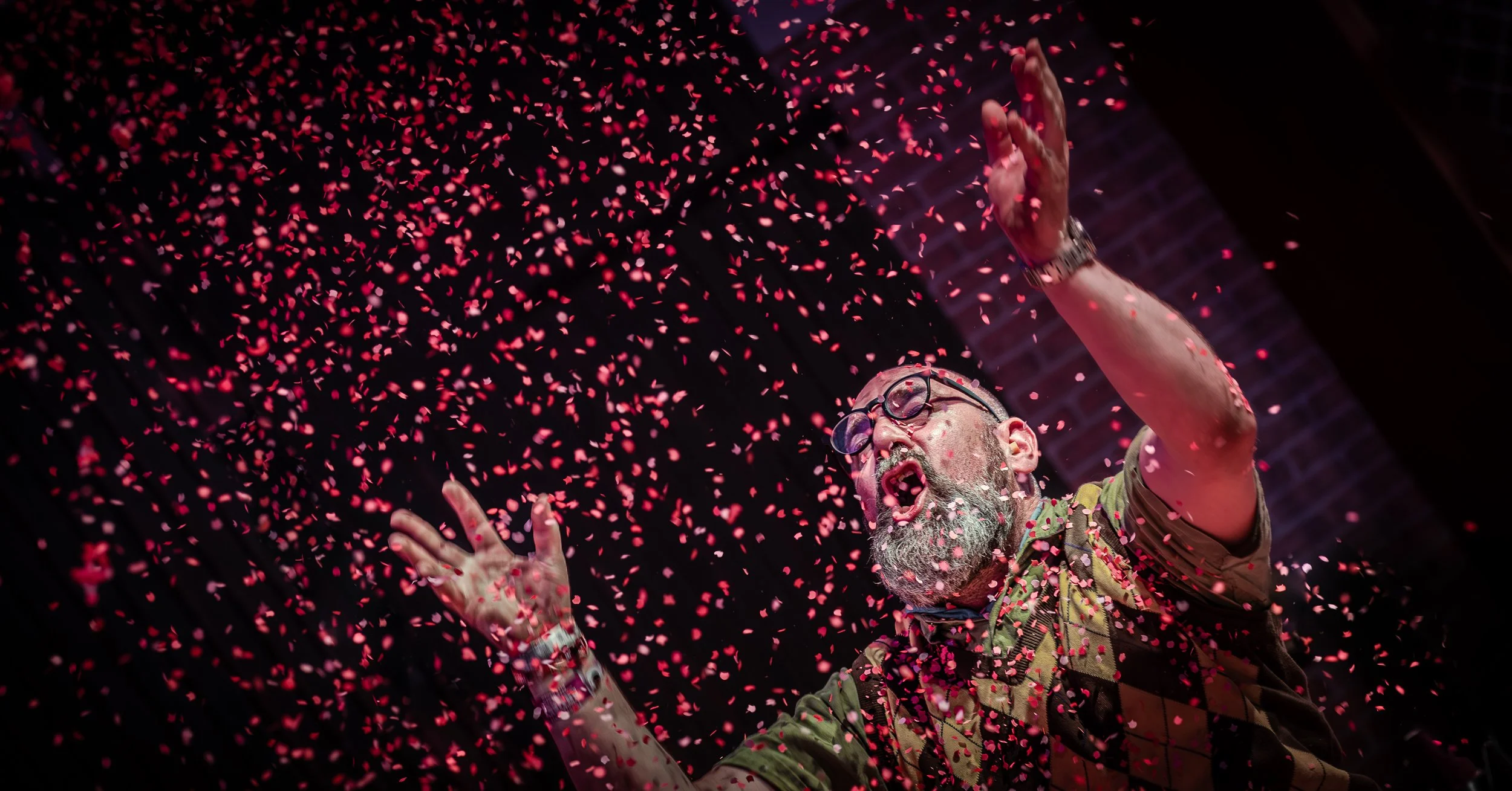 Man met glazen, grijs haar en een baard premierend met roze confetti mid-performance op een donkere achtergrond.