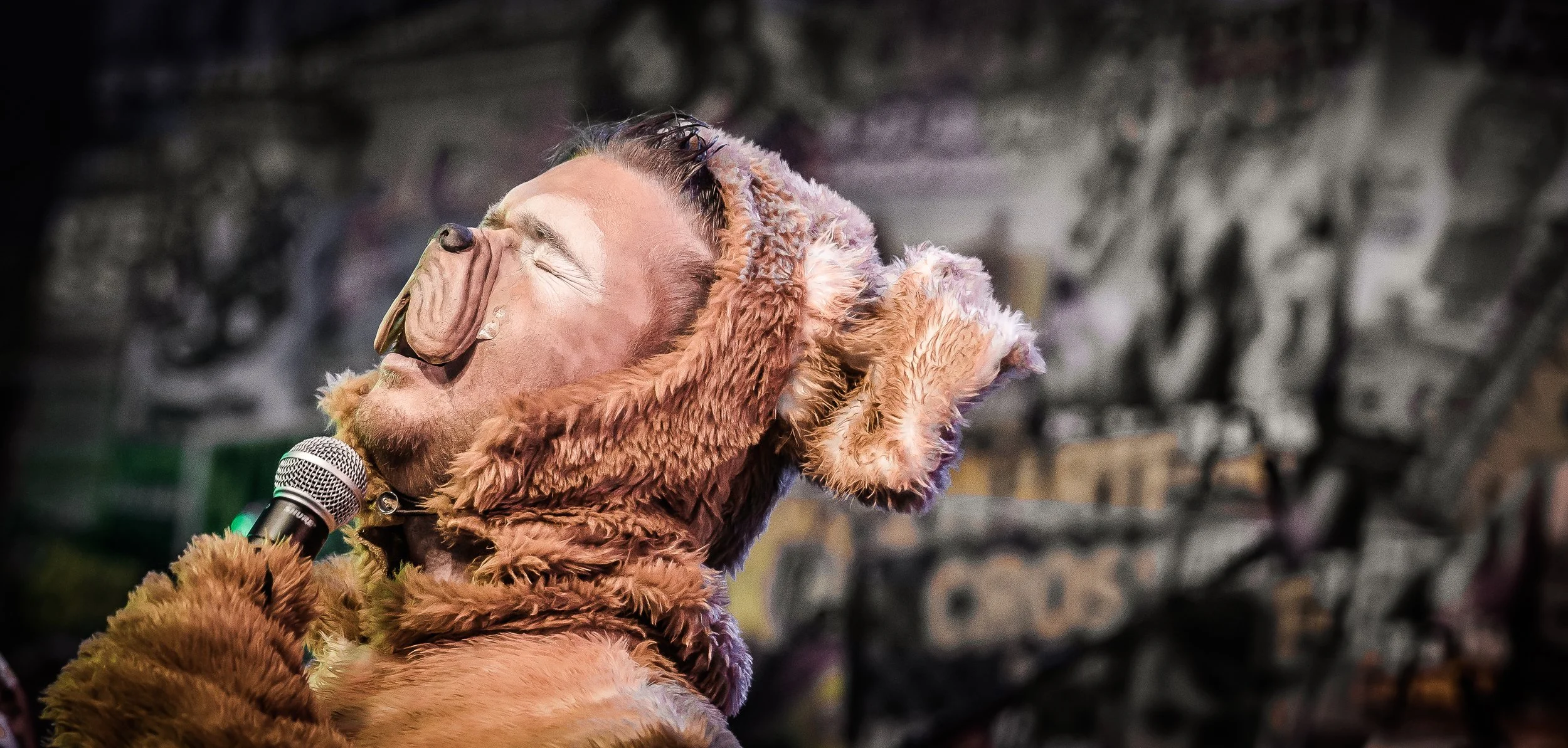 Man met een hondenkostuum met grote oren, lachend en met gesloten ogen, houder van een microfoon in een kamer met graffiti en posters op de muur.