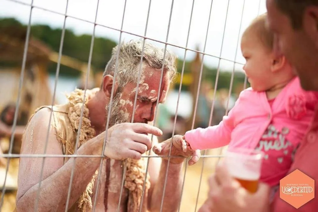 Man met schmink op zijn gezicht achter een ijzeren hek, terwijl hij een baby met een pink aanraking aanraakt.