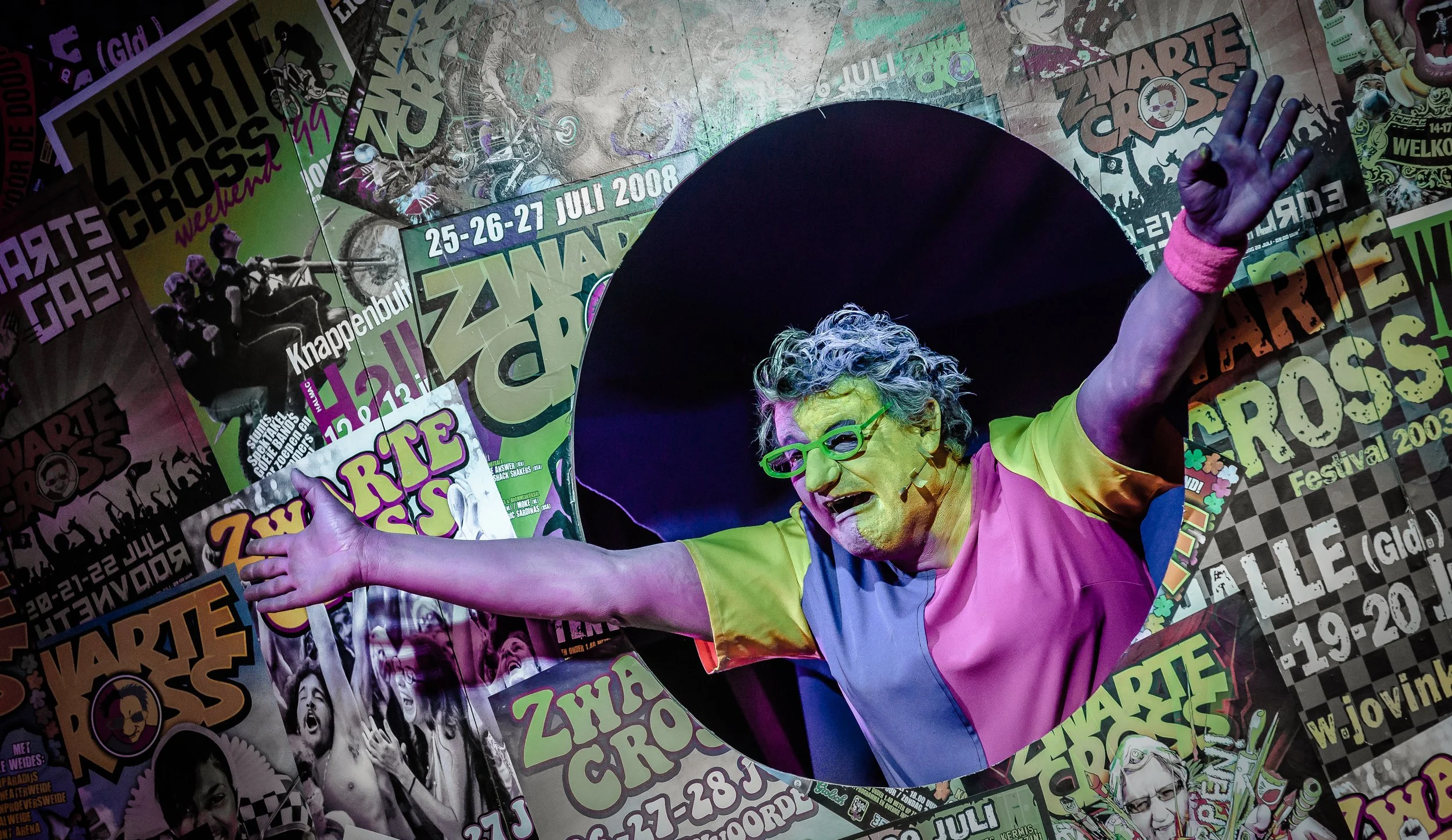 Het Zwarte Cross logo met verrassend gekleurd haar, groen bril, roze en gele kleding, uitgestrekte armen, omringd door posters van Zwarte Cross festival.