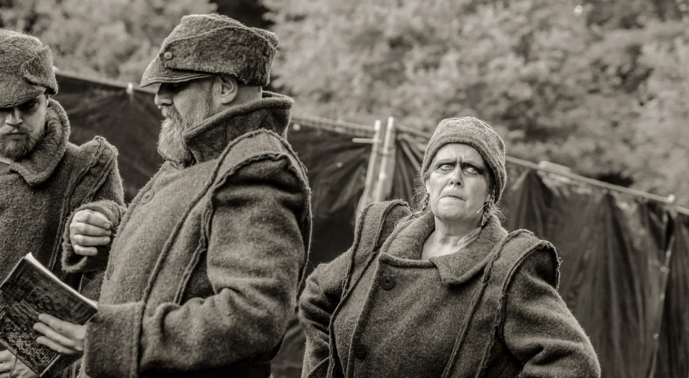 Drie mensen in historische militaire kleding met mutsen, twee mannen en een vrouw, buiten met een tent op de achtergrond.