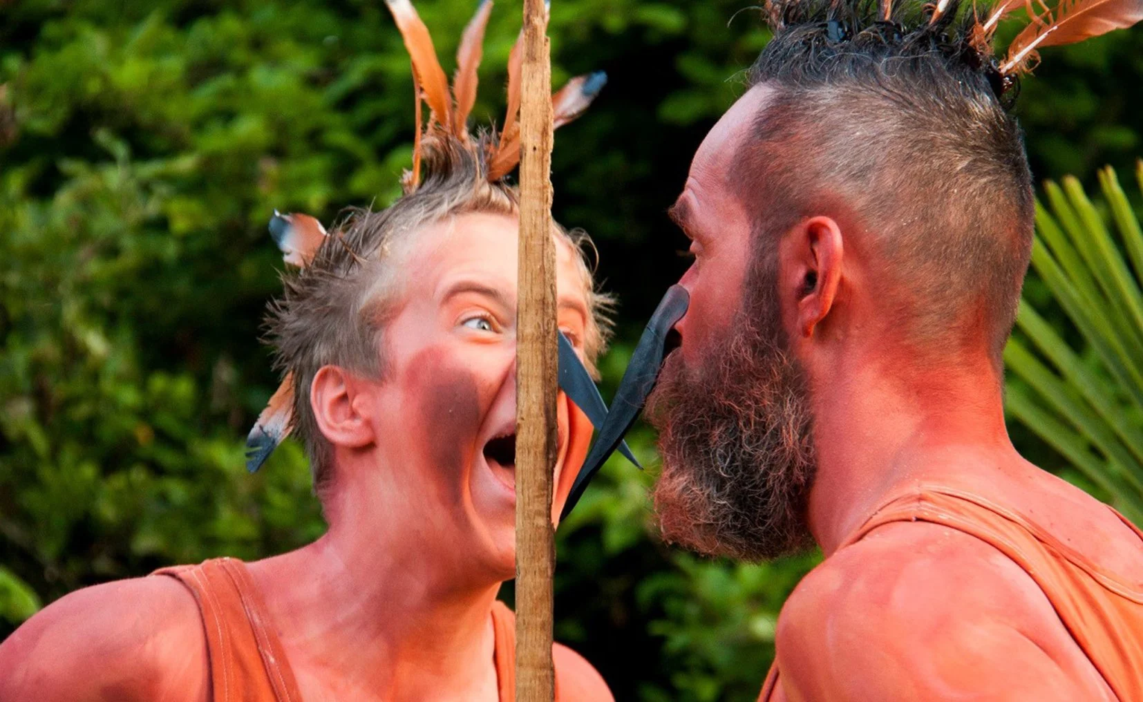 Twee mensen met gezichtsschildering en veren in het haar, in een omgeving met groen gebladerte.