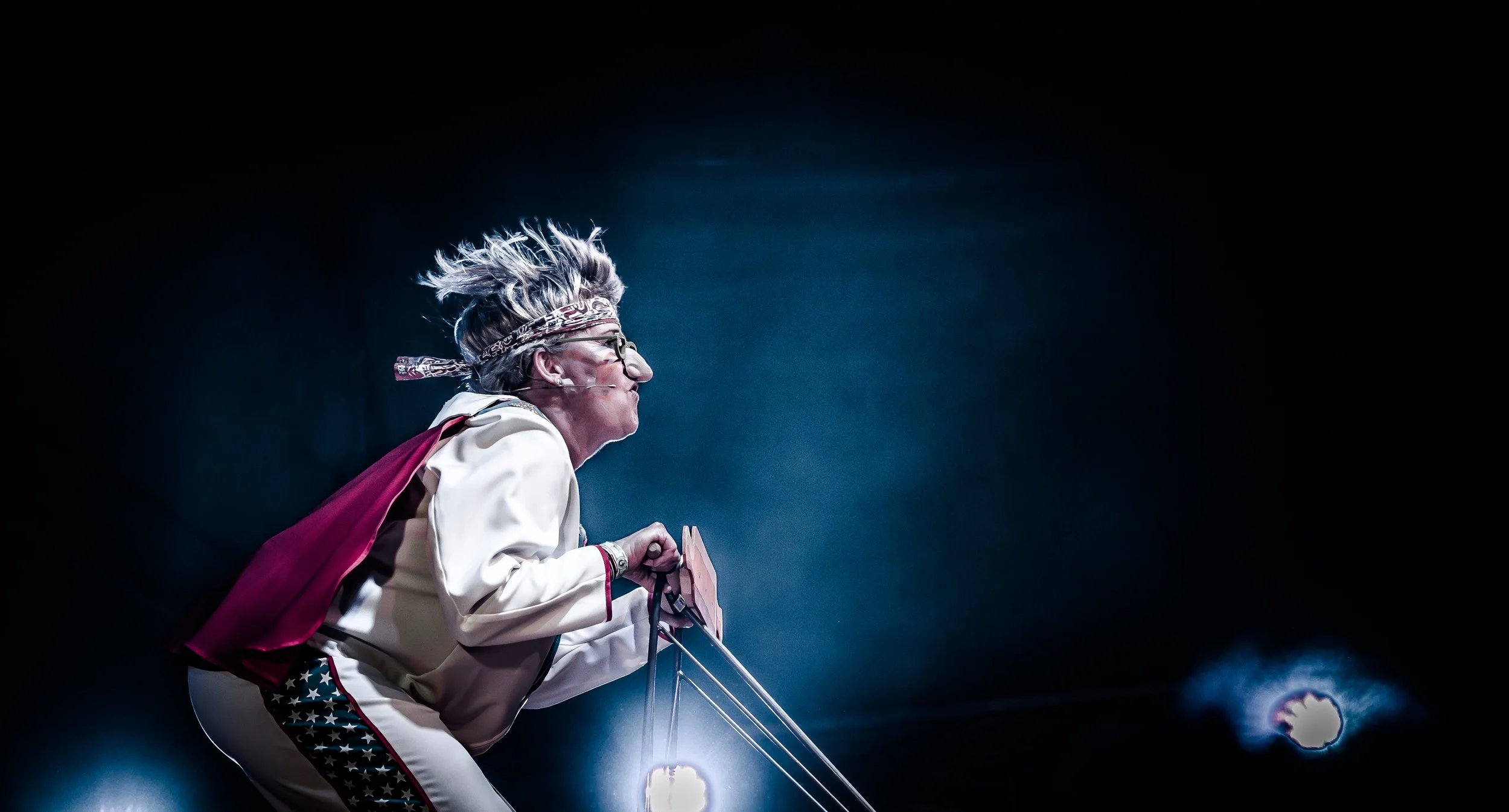 Een persoon in een kostuum met een cape, gekleed als een superheld, staat op een podium met felle lichten die op haar gericht zijn.