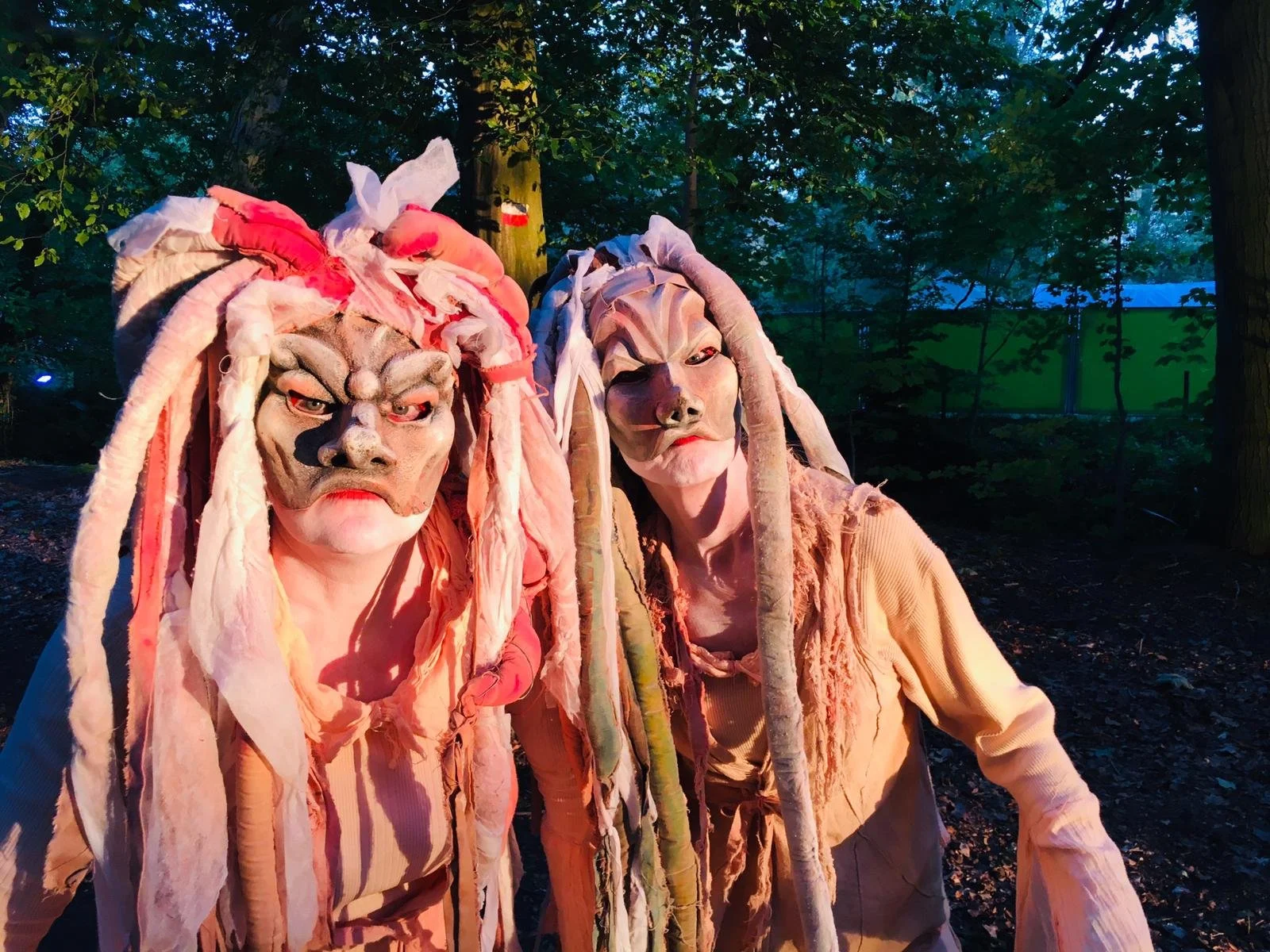 Twee mensen in uitgebreide maskers en kostuums die lijken op fantasywezens in een bos, mogelijk voor een evenement of performance.