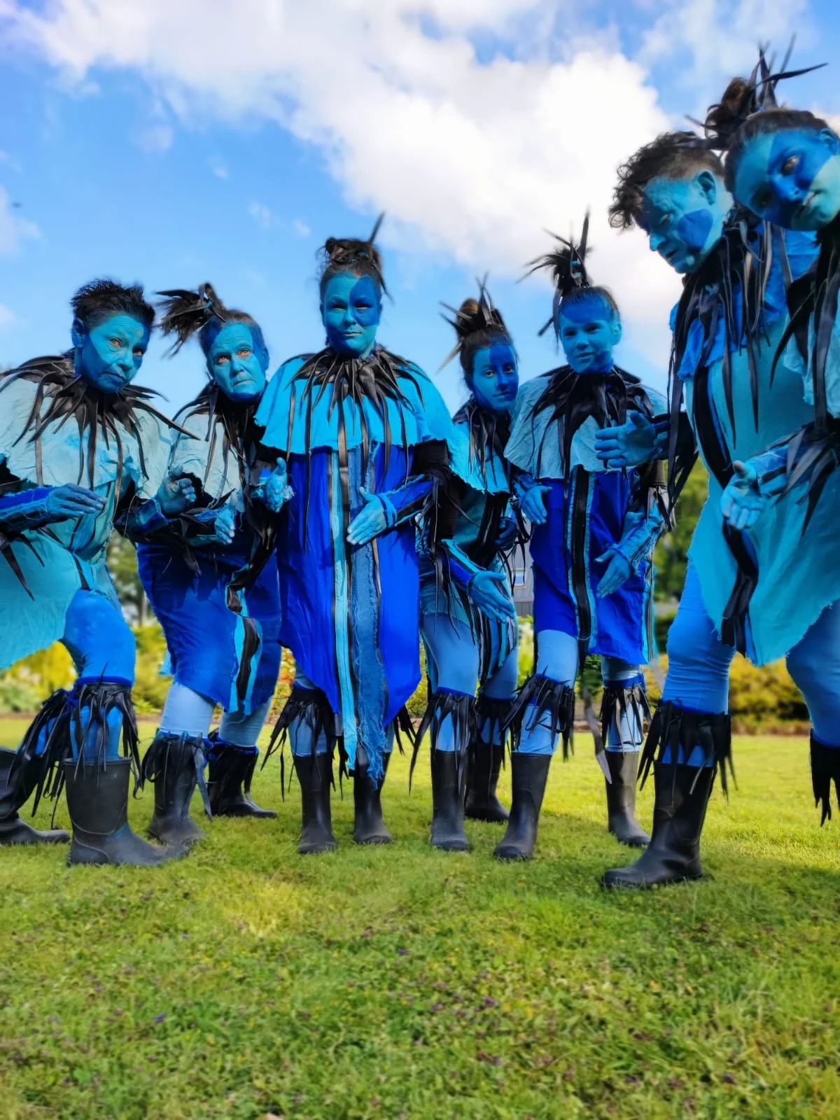 Mensen in blauw geklede en gemaskerde kostuums met blauwe gezichten en zwart getekende details, mogelijk voor een festival of evenement, staand op gras met een blauwe lucht en wolken op de achtergrond.