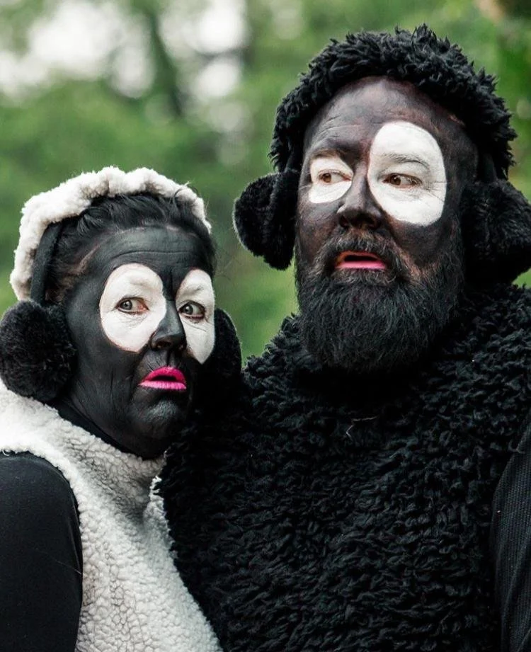 Twee mensen in zwart-wit gezichtsschmink en kostuums, gekleed als schapen, in een bosomgeving.