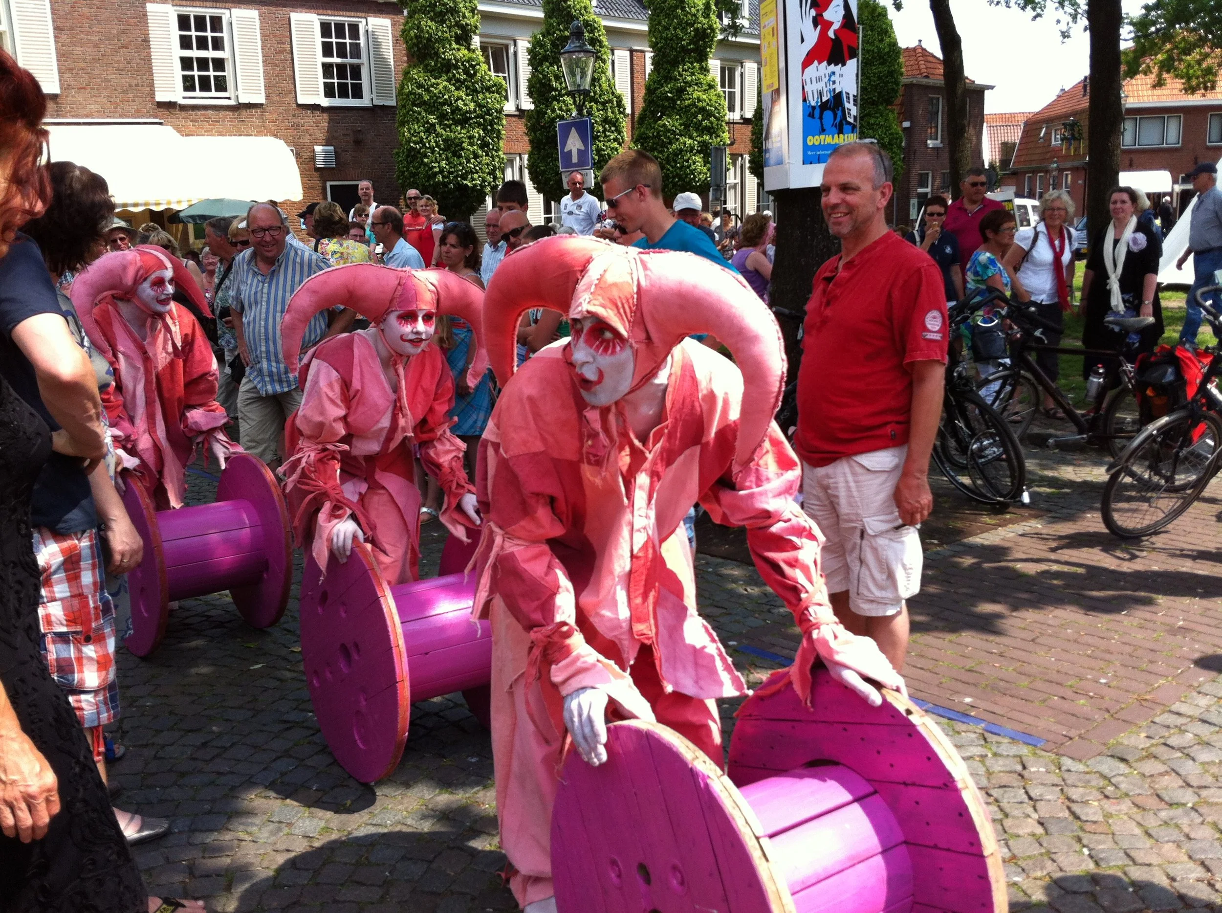 Narren in roze kostuums met wit geschminkte gezichten en hoed-achtige pruiken, waarbij ze grote roze en paarse katrollen voortduwen, omringd door een publiek op een straat in een stadsdeel.