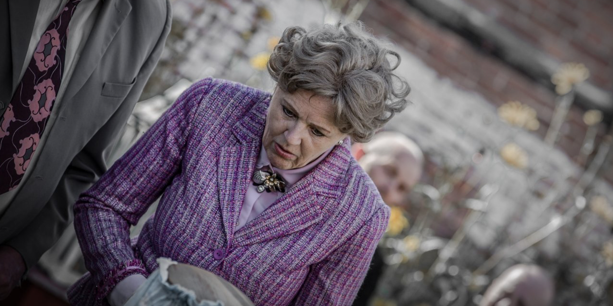 Oudere vrouw met grijs, krullend haar draagt een paarse bloemenbroche en een geruite roze paarse jas, kijkt geboeid naar iets in haar hand, omgeven door andere mensen in een tuin.