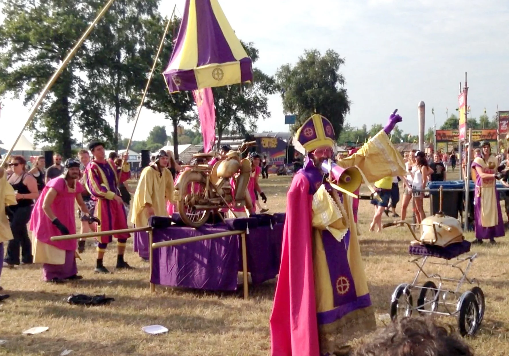 Een kleurrijke straatartiest in een theatrale kostuum met een masker en een hoed, die een publiek entertaint met een blaaskunst en een fantasievolle, felle outfit die roze en geel combineert, op een festivalterrein met mensen en tenten op de achtergrond.