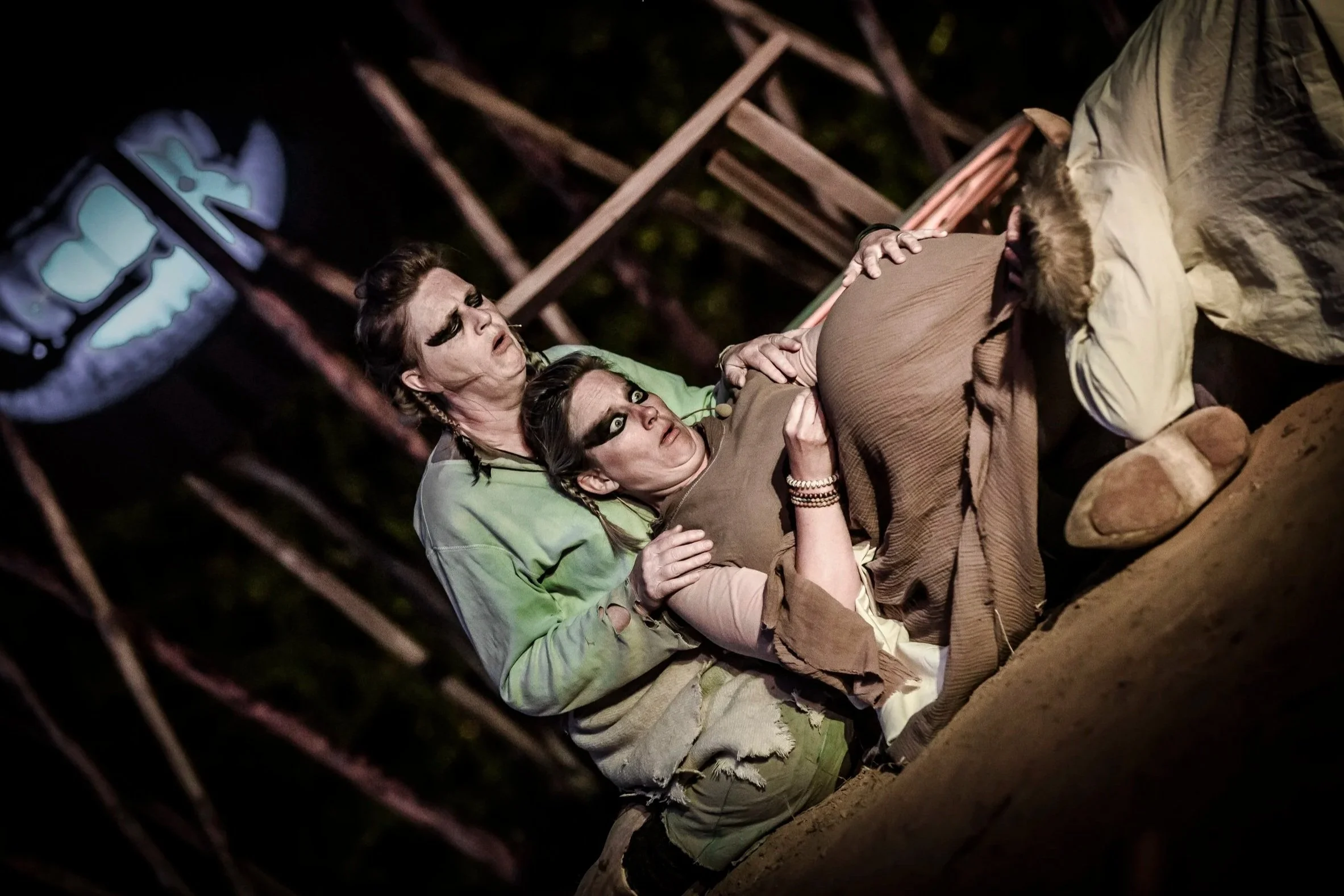 Theater scène met twee vrouwen in dramatische pose, een ligt op de grond met een zwangere buik en de ander houdt haar vast, donker decor op de achtergrond.