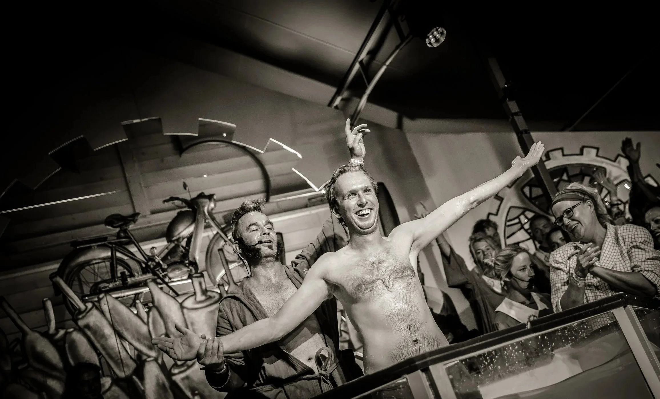 Een blije man zonder overhemd in een voorstelling, omringd door mensen die lachen, met een decor van grote tandwielen op de achtergrond.