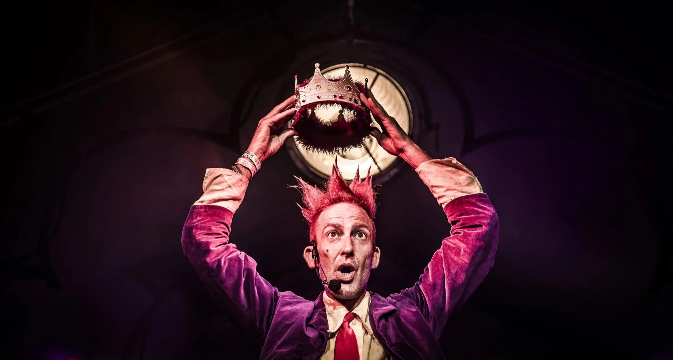 Man met felrode haar en een verwarde uitdrukking houdt een kroon boven zijn hoofd in een donkere ruimte met een licht van boven.