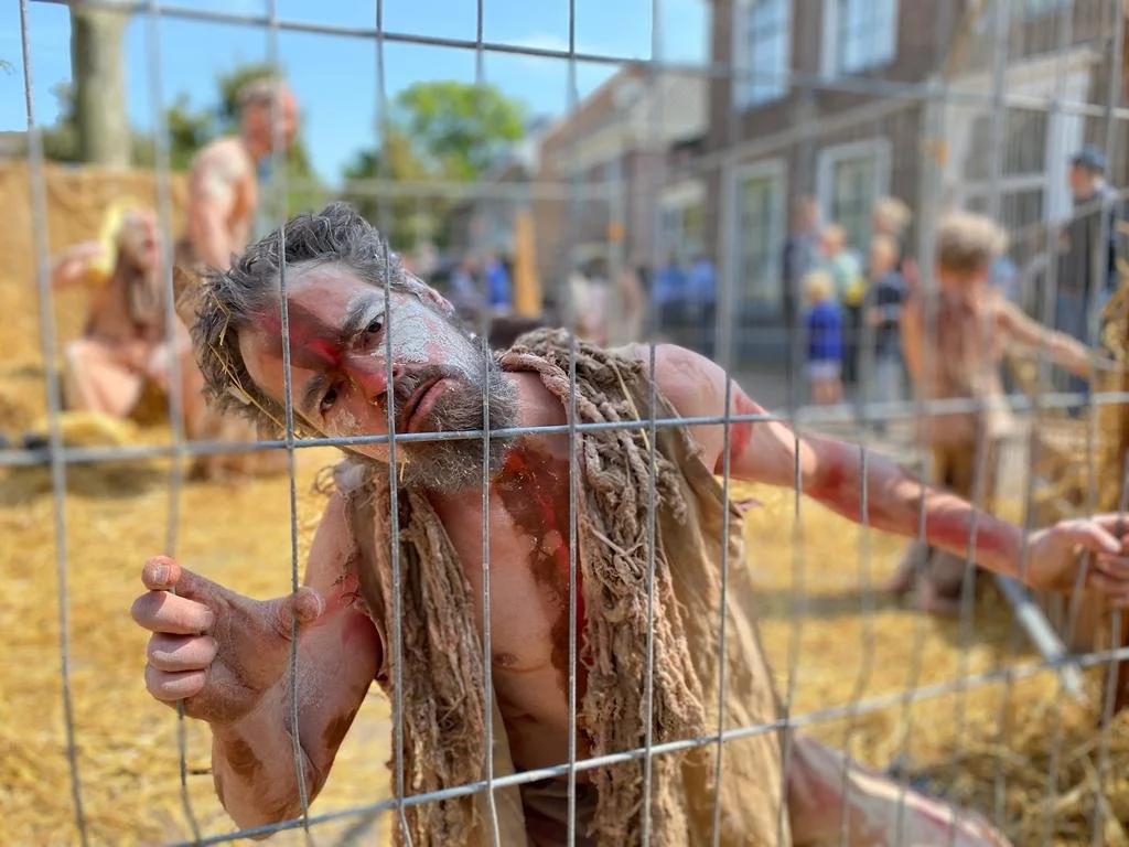 Een man met schmink achter een metalen hek, met oermenschen op de achtergrond.