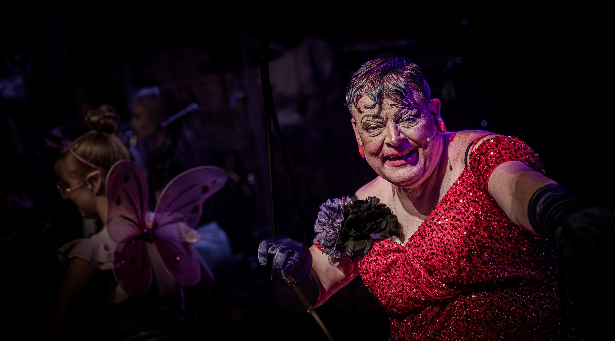 Persoon in een glitterende rode jurk met make-up en haar in een stijlvol kapsel, met een grote bloem op de jurk, houdt een stok en poseert theatrale op een donkere achtergrond.