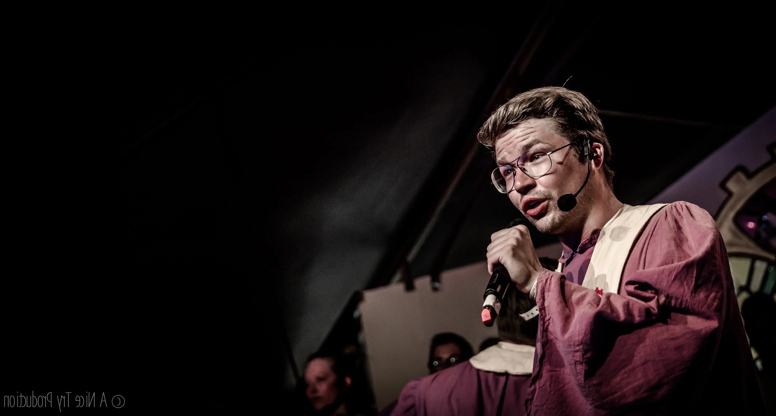 Man met bril en microfoon geeft opvoering, met drie mensen op de achtergrond.