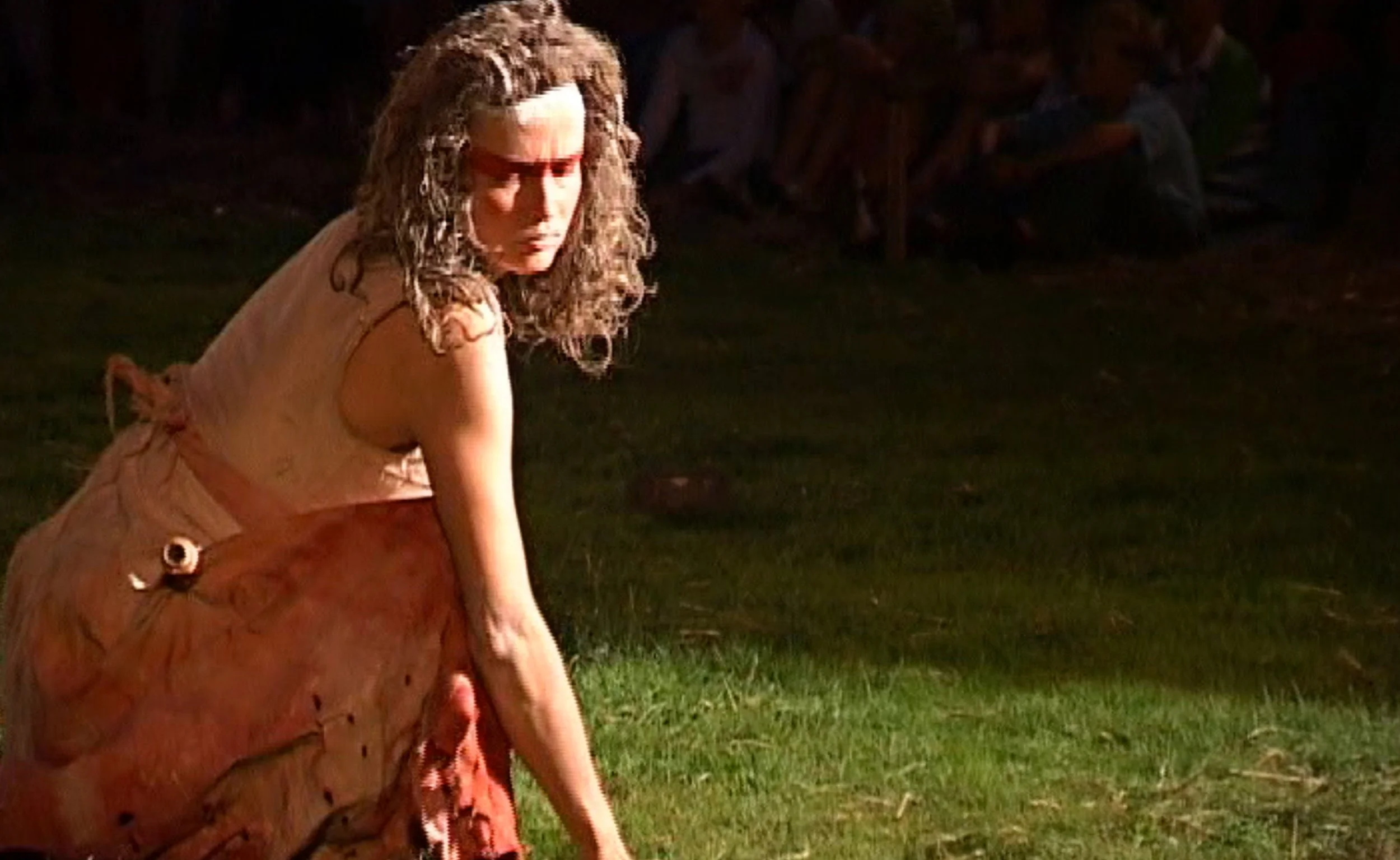 Een vrouw met lang, gekruld haar in een setting met gras en donkere achtergrond.