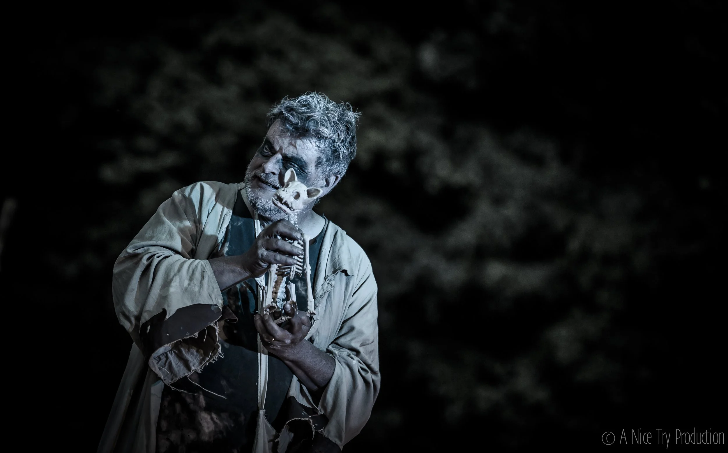 Een man met grijs haar en een baard houdt een schedel van een dier en een pop van een dier, beide in zijn handen, tegen een donkere achtergrond.