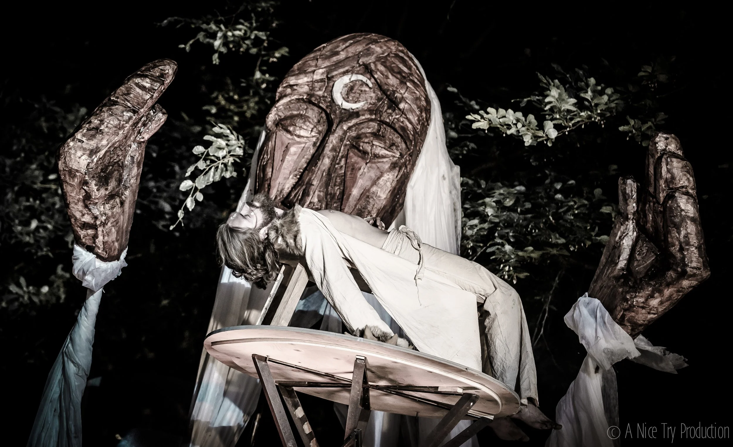 Het beeld toont een nachtelijke scène met een sculptuur van een man die op een kruis ligt, omringd door grote houten handen met witte stoffen banden. De achtergrond bevat donkere bladeren.