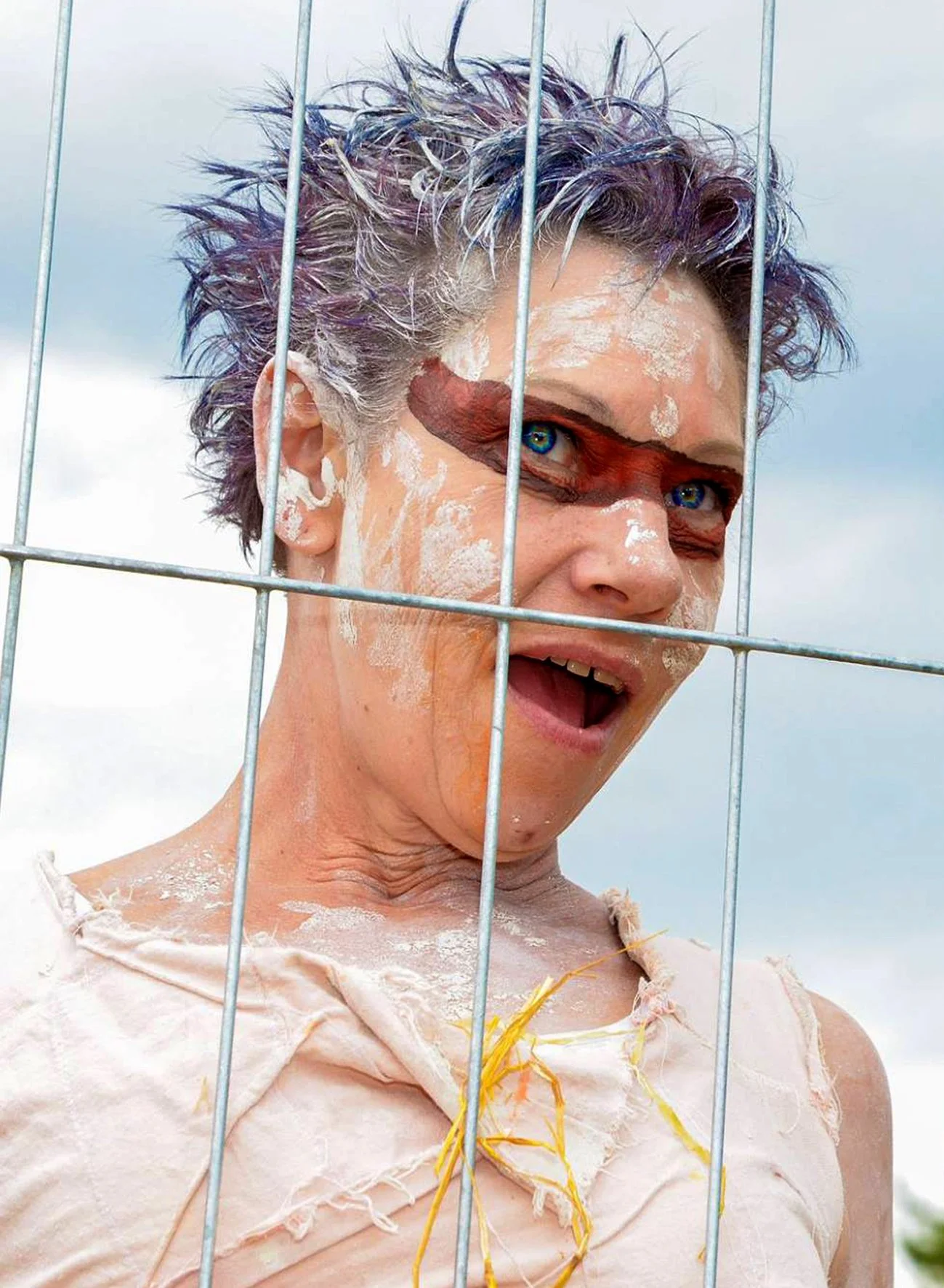 Een vrouw met afgekleurde, ruige haar en opvallende gezichtsmake-up, die door een metalen hek kijkt.