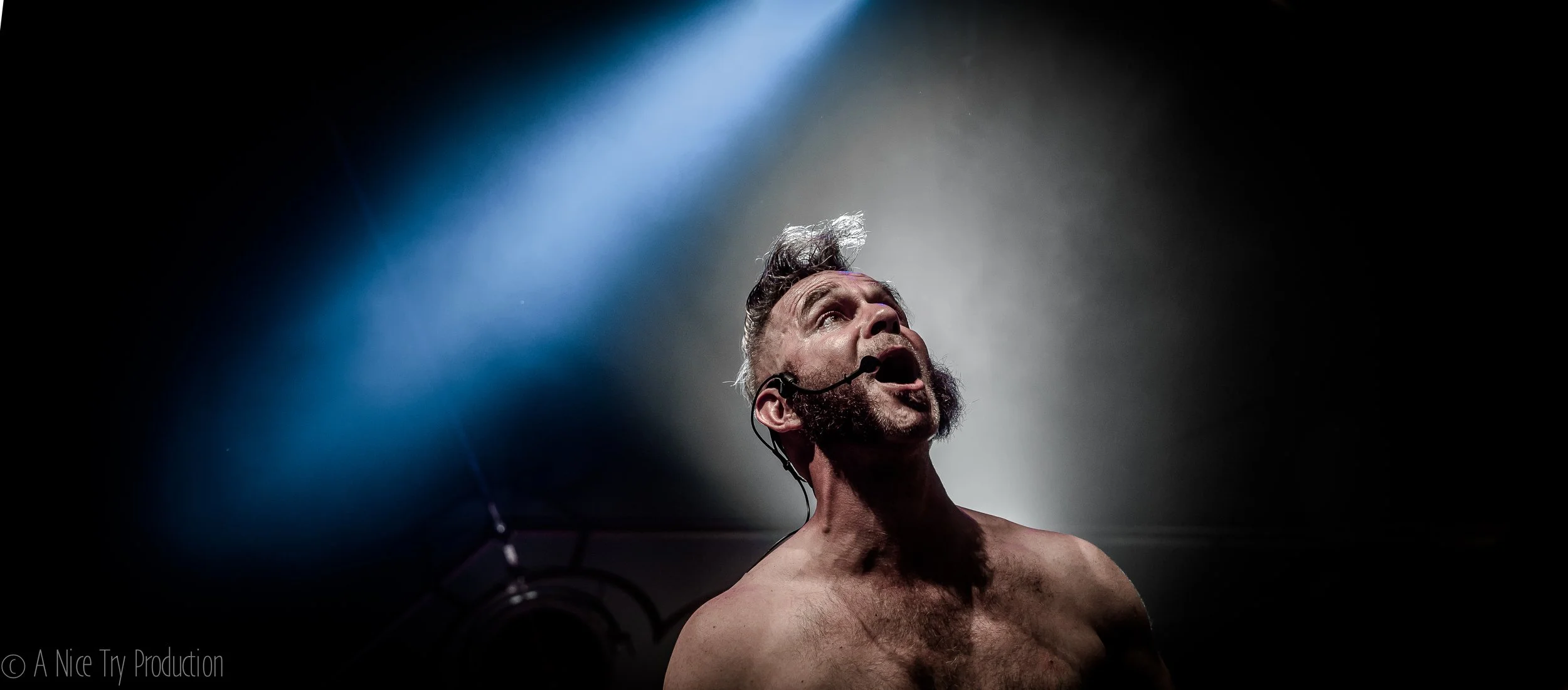 Man met een microfoon op een podium, verlichte met een witte spotlight, met een blote bovenlichaam.