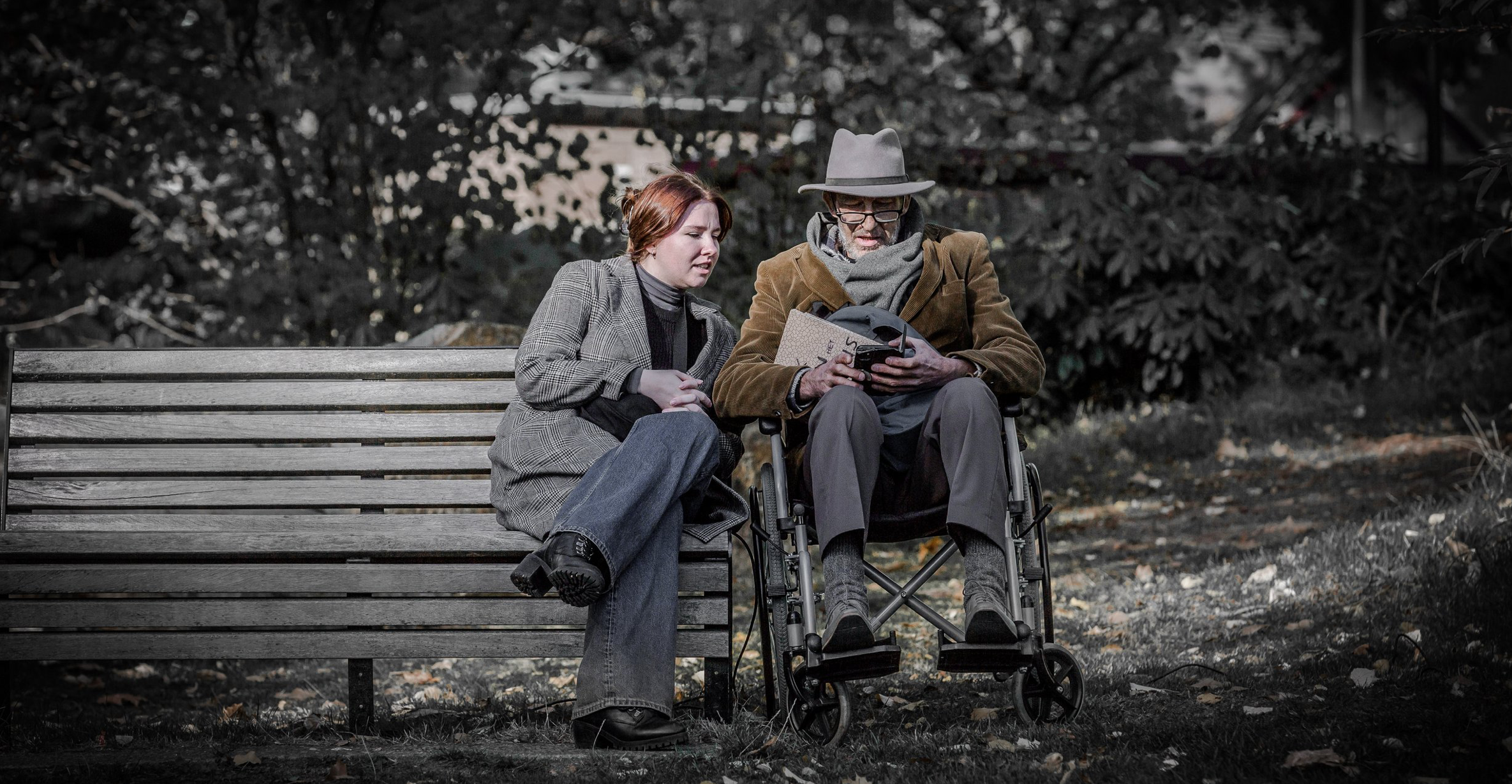 Twee mensen zitten op een bank in een park, een vrouw en een man in een rolstoel, verdiept in een boek of tablet. De achtergrond bestaat uit bomen met herfstbladeren op de grond.