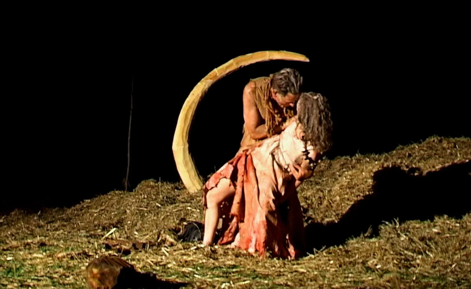 Twee acteurs op een podium, een man die een vrouw vasthoudt, met een boog in de achtergrond, op een podium met aarde of zand als vloer.